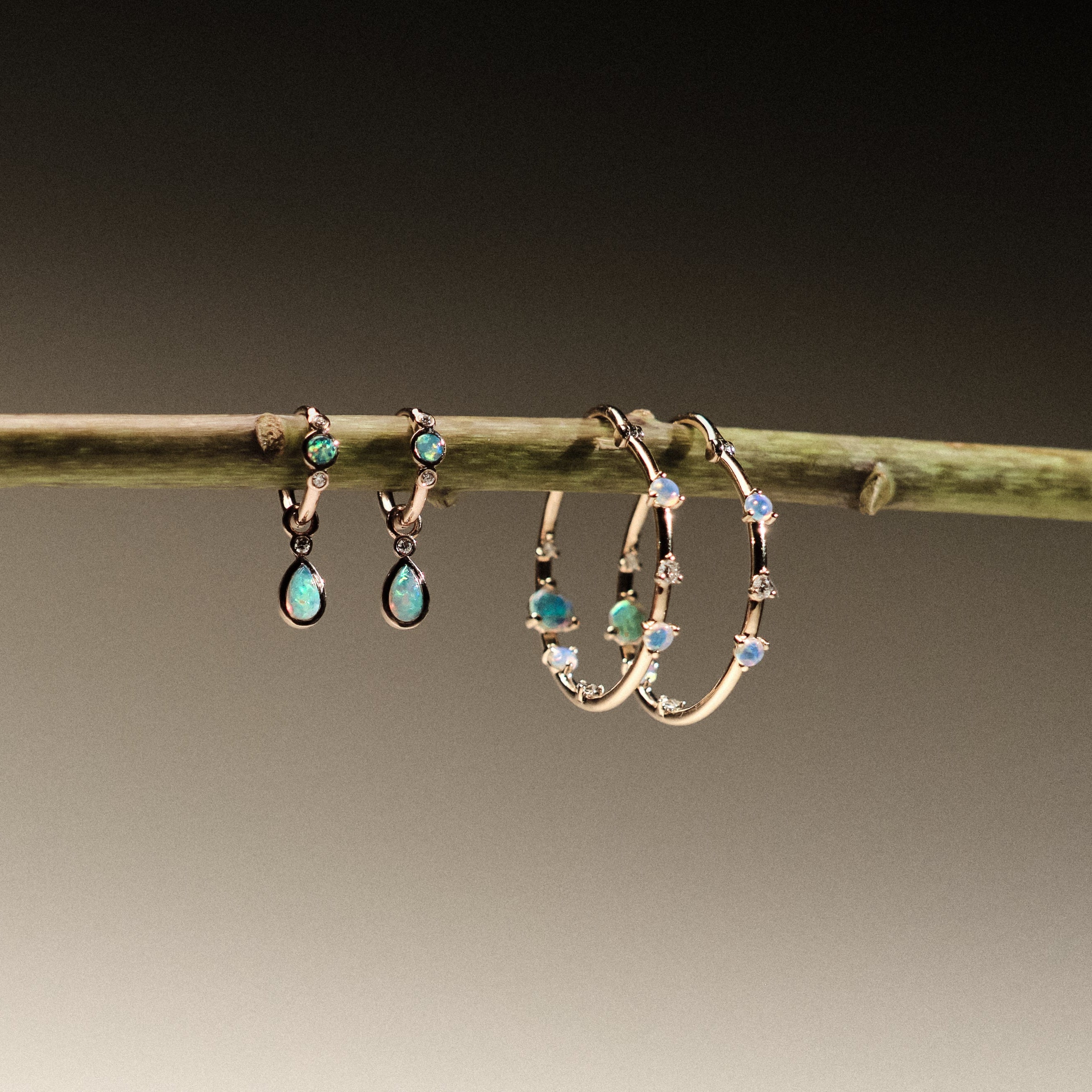 Two pairs of earrings made of 14k gold, lab diamonds and australian opals, hanging on a branch against a neutral background