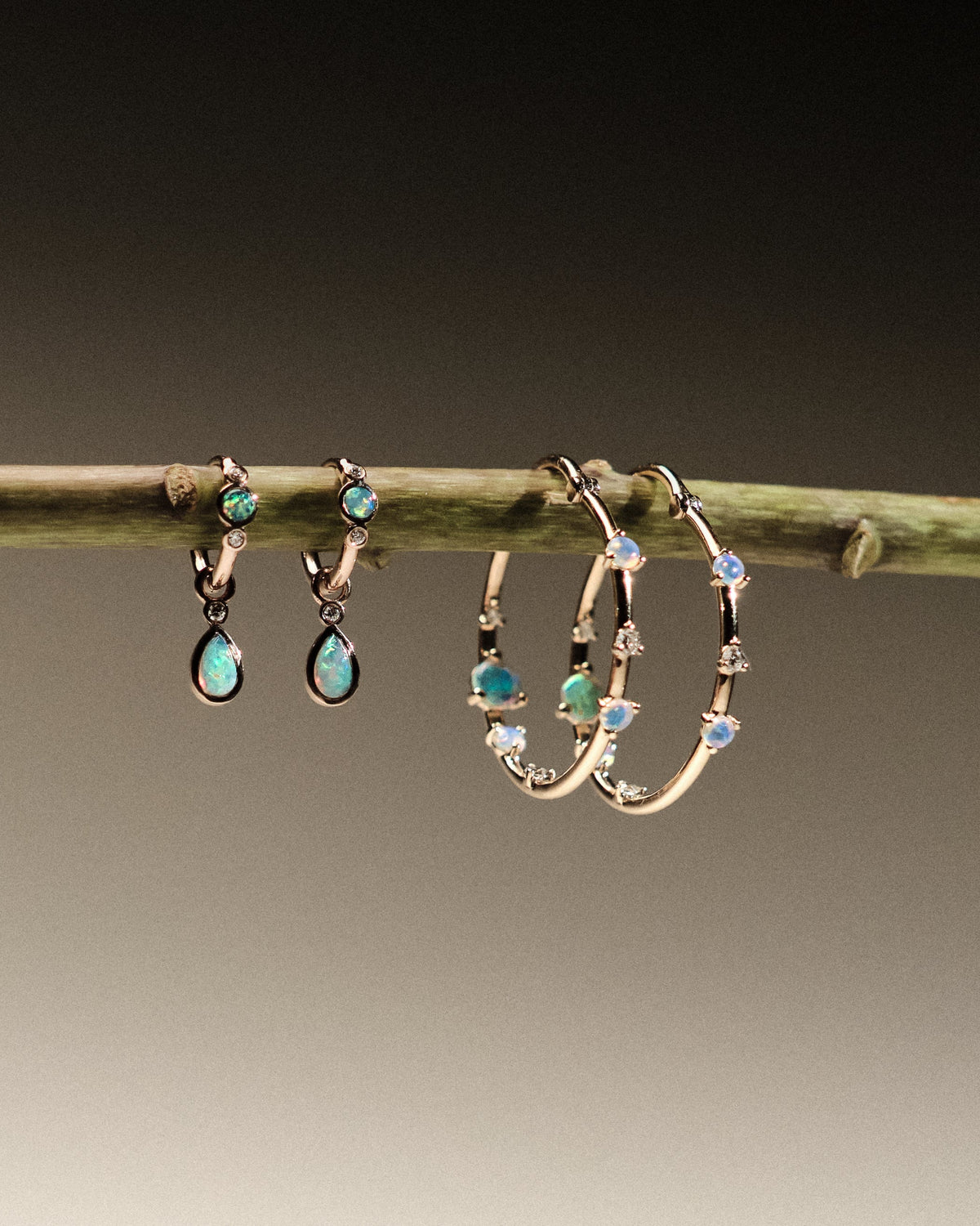 Two pairs of earrings made of 14k gold, lab diamonds and australian opals, hanging on a branch against a neutral background