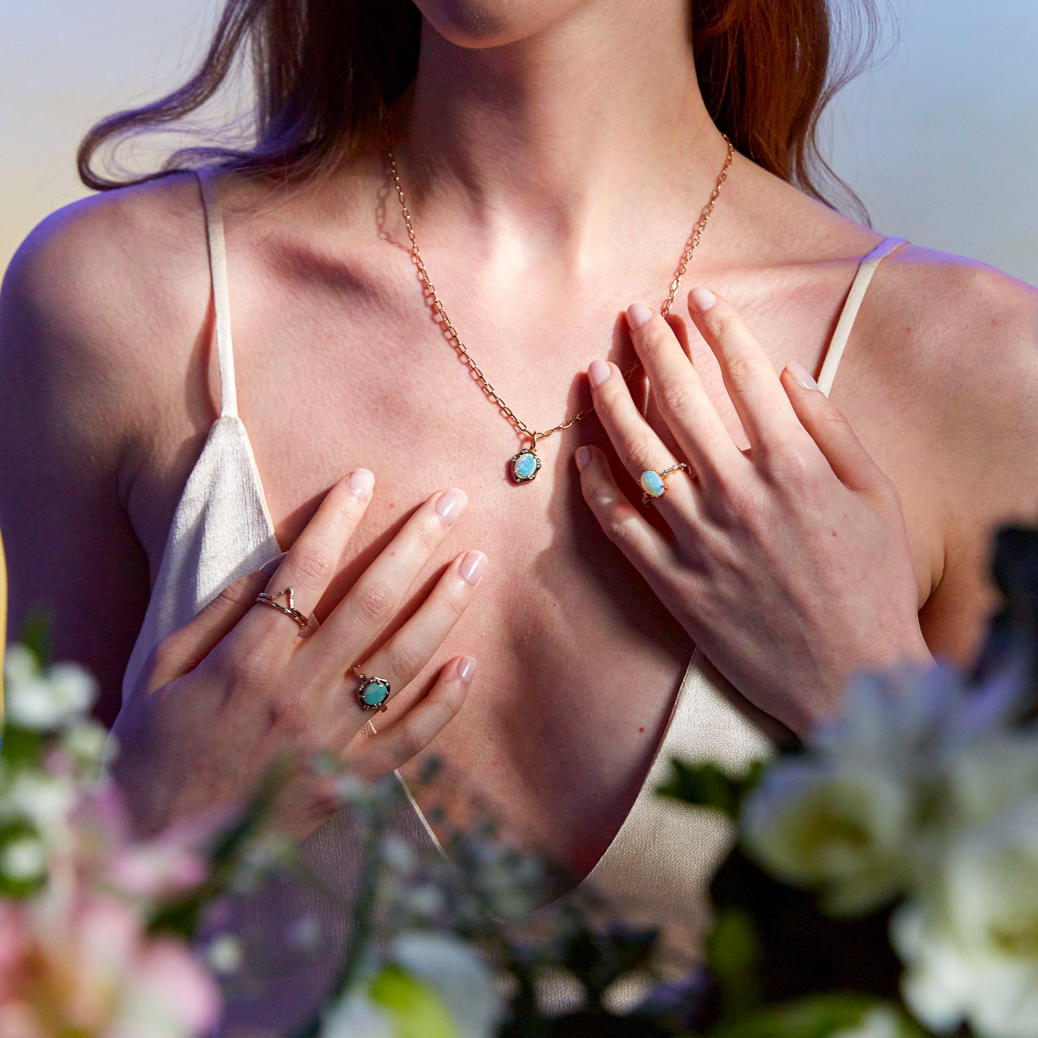 Hands showcasing gold rings and a pendant, with an opal center stones and white diamond accents on the band, placed on a floral background.