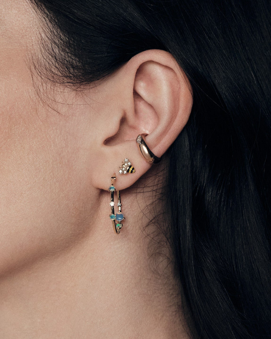 Close-up of a person wearing gold hoop earrings with diamonds and australian opals, baby bee enamela and diamond studs and marquise diamond earcuff.