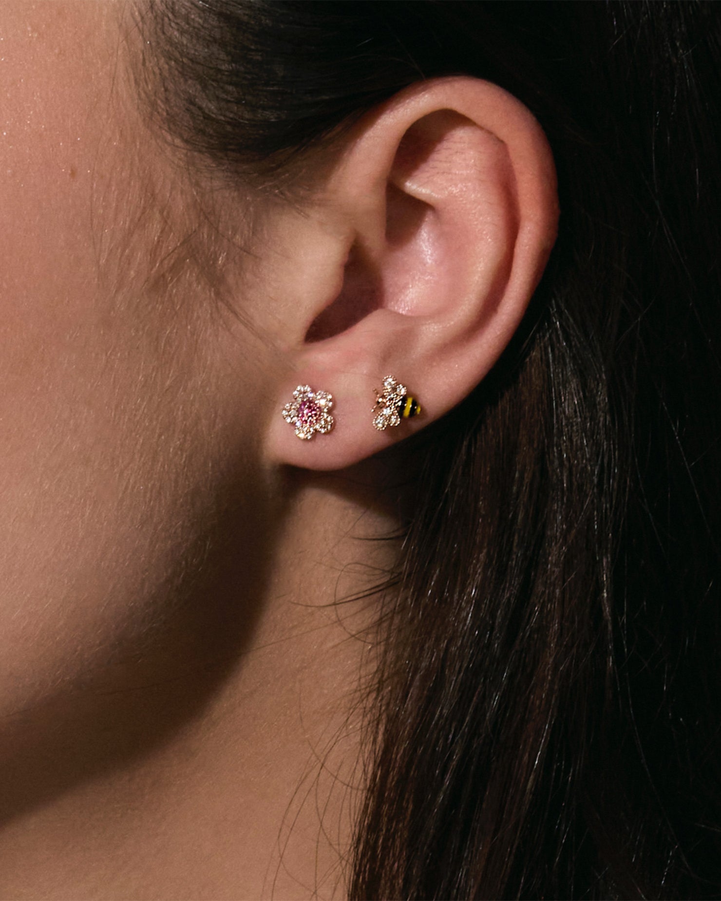 Close-up of an ear wearing two earrings , one is a gold bee with enamel black and yellow stripes and the other one is a diamonds and rubies cherry blossom flower