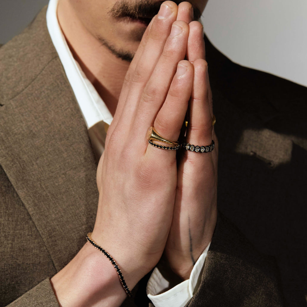 Close-up of a person's hands with a black diamonds tennis ring, black gold and white diamonds tennis ring, signet gold ring and a black diamonds tennis bracelet, wearing a suit.