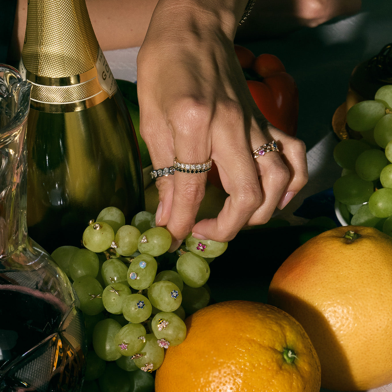 Close-up of a hand with eternity diamonds rings picking grapes among fruits and a bottle, possibly champagne.