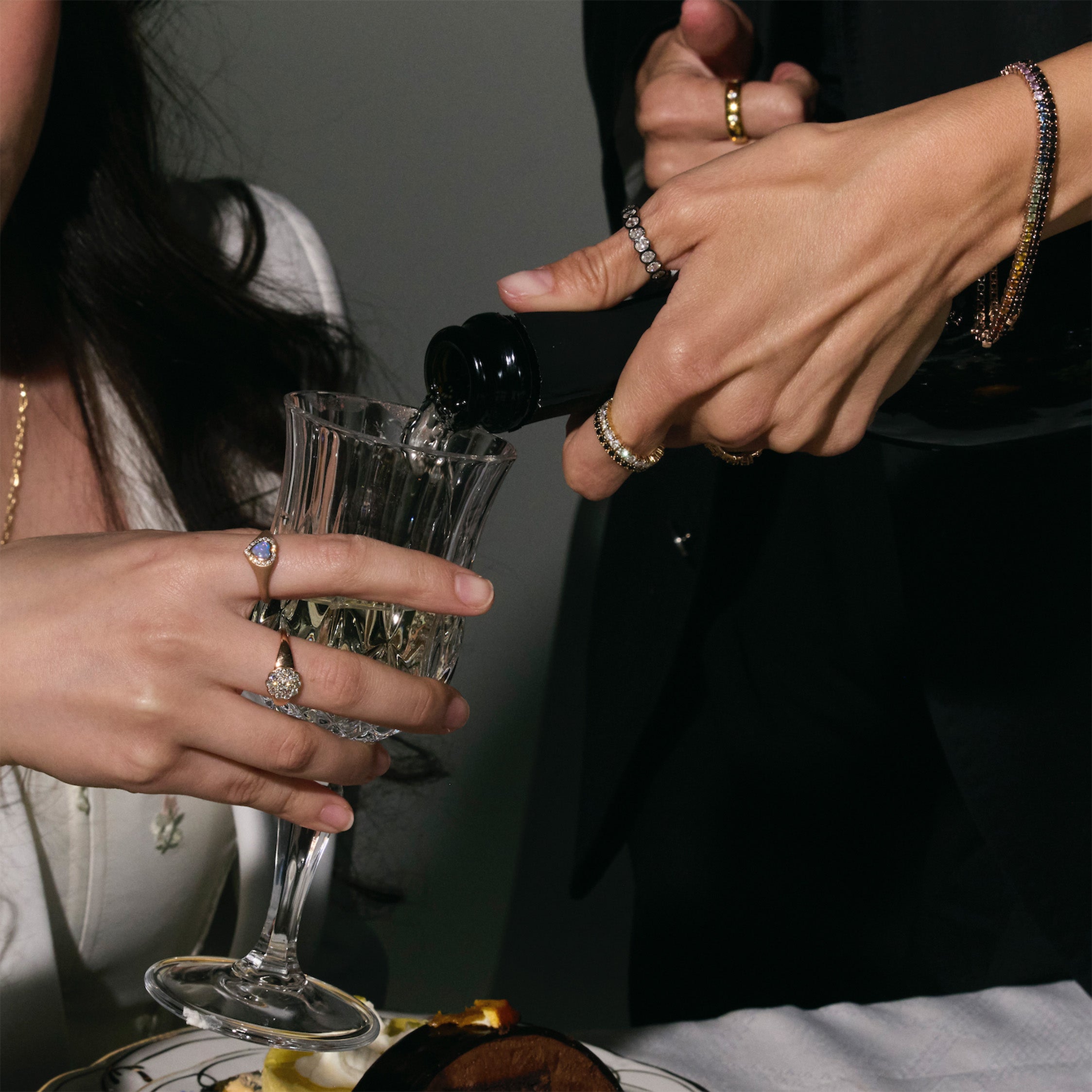 a woman pouring a glass of champagne to another one, both of them are wearing exclusive gold jewelry, eternity rings with white and black diamonds, eternity tennis bracelets