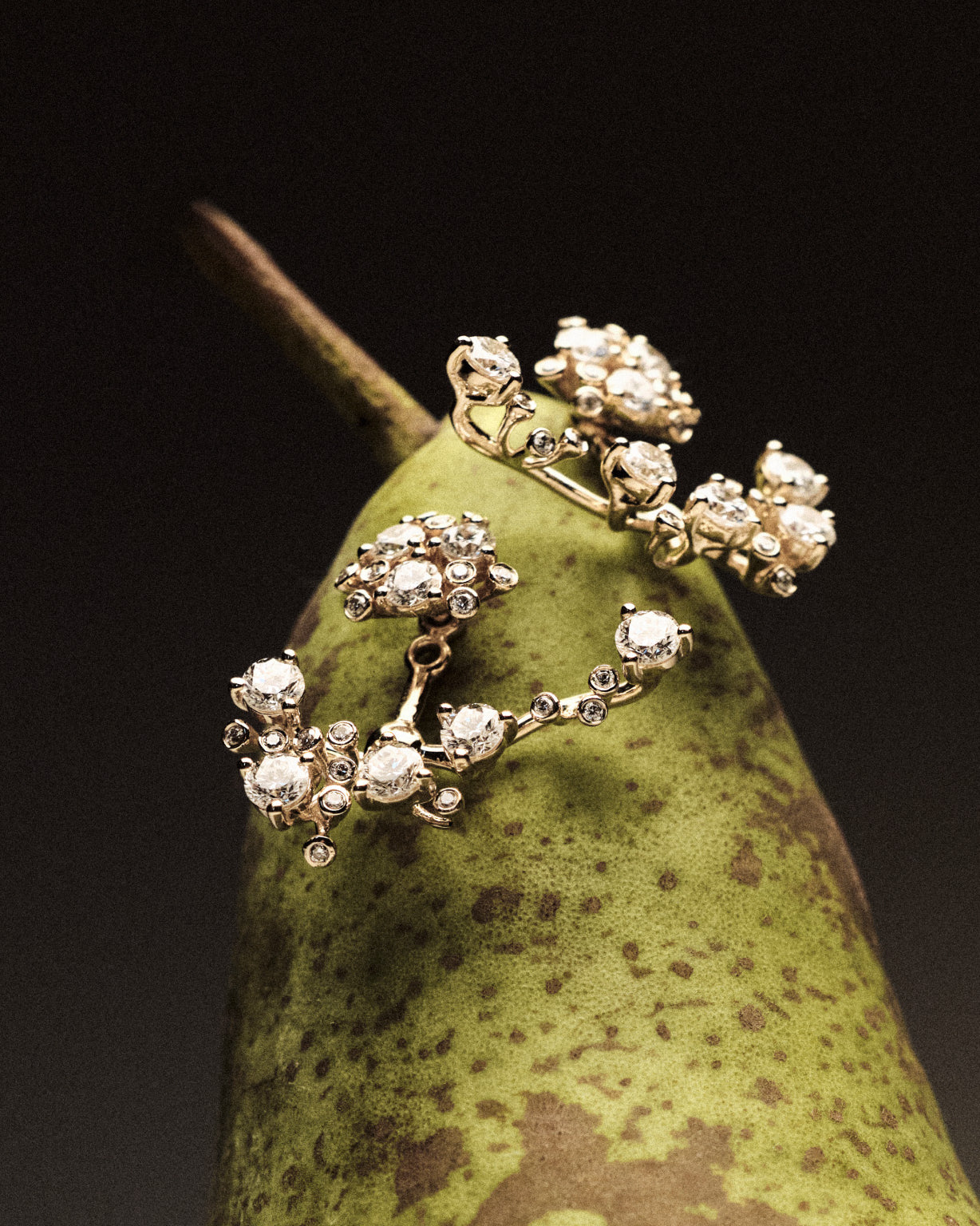 Gold and diamond earrings on a green pear against a dark background.