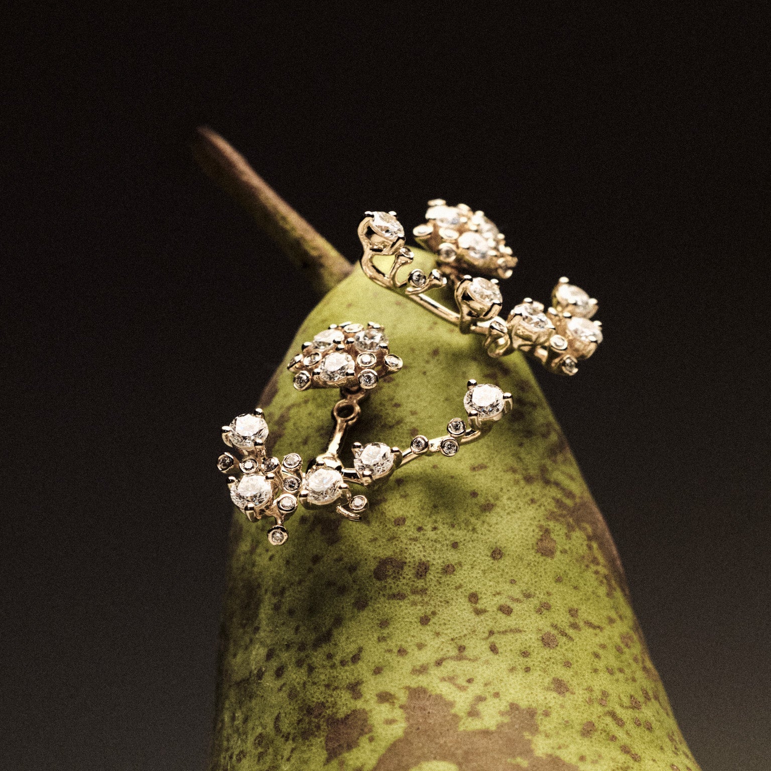 Gold and diamond earrings on a green pear against a dark background.