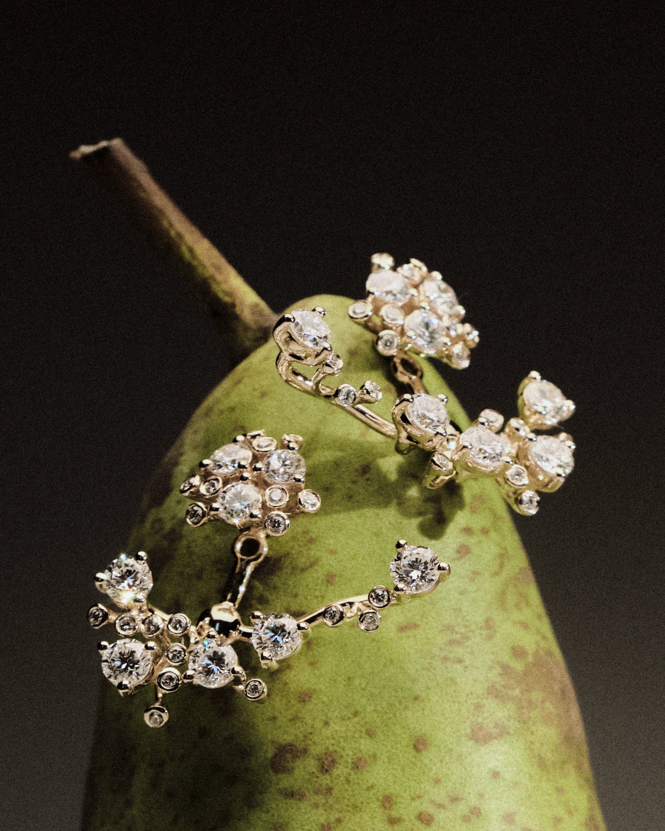 Gold and diamond earrings on a green pear against a dark background.