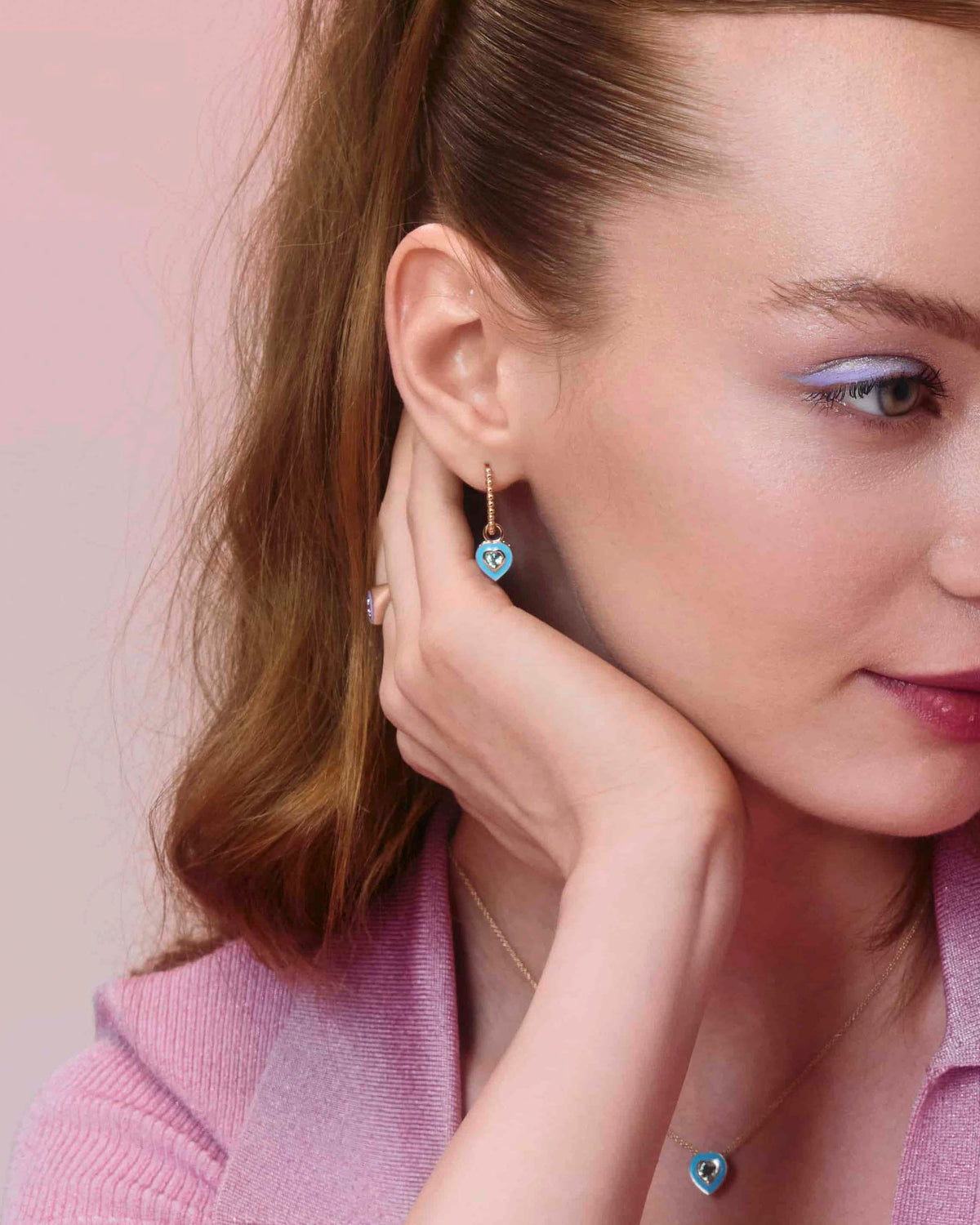 gold hoop earring with a ball design, with enamel heart accessory, on model's ear displayed against a pink background.
