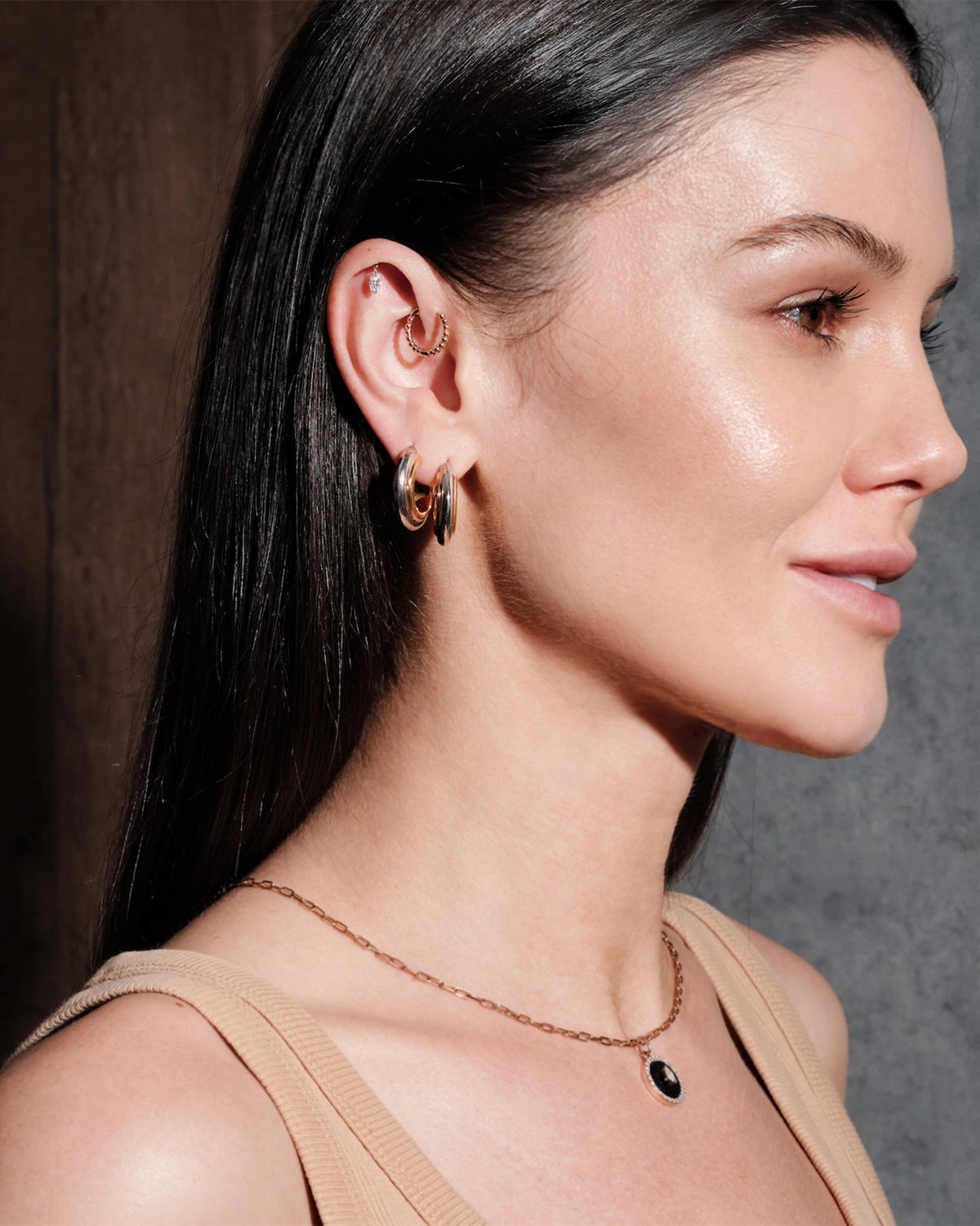 Woman wearing multiple hoop earrings, thick gold hoops, floating marquise diamond stud, simple beaded hoop, with a dark background