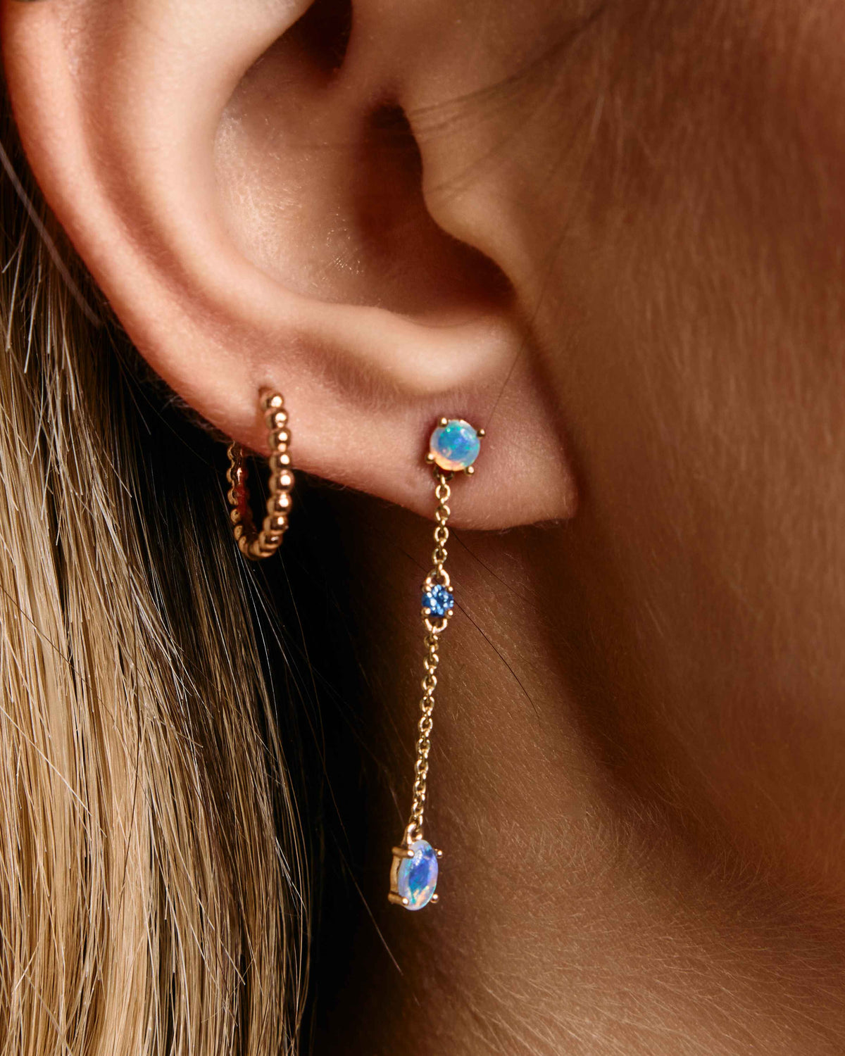 A close-up image of a woman's ear wearing a rose gold, chain link earring with an Australian Opal and a green sapphire.