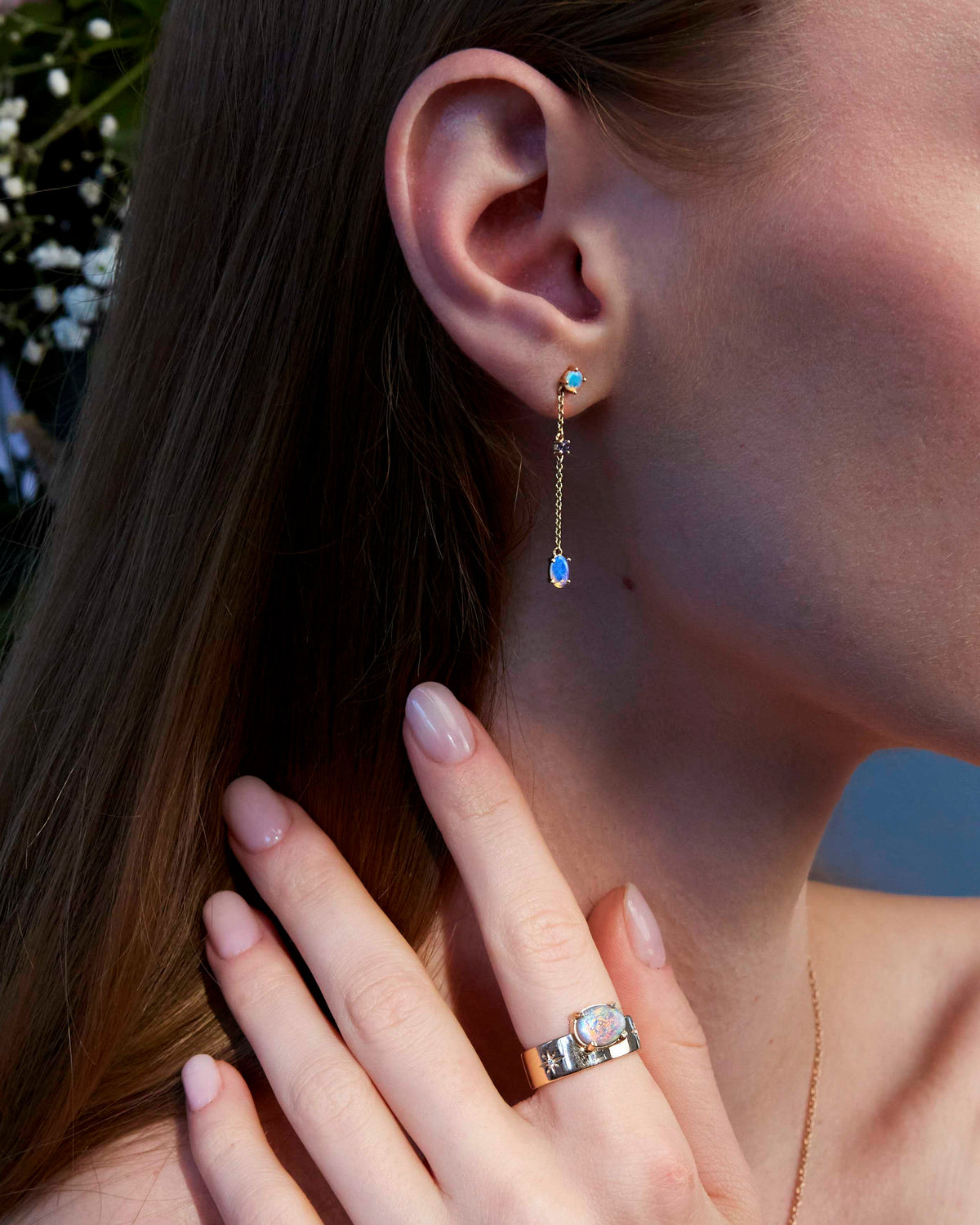 A person wearing a pair of gold earrings with round blue or green sapphires and Australian Opals, standing near a floral backdrop.