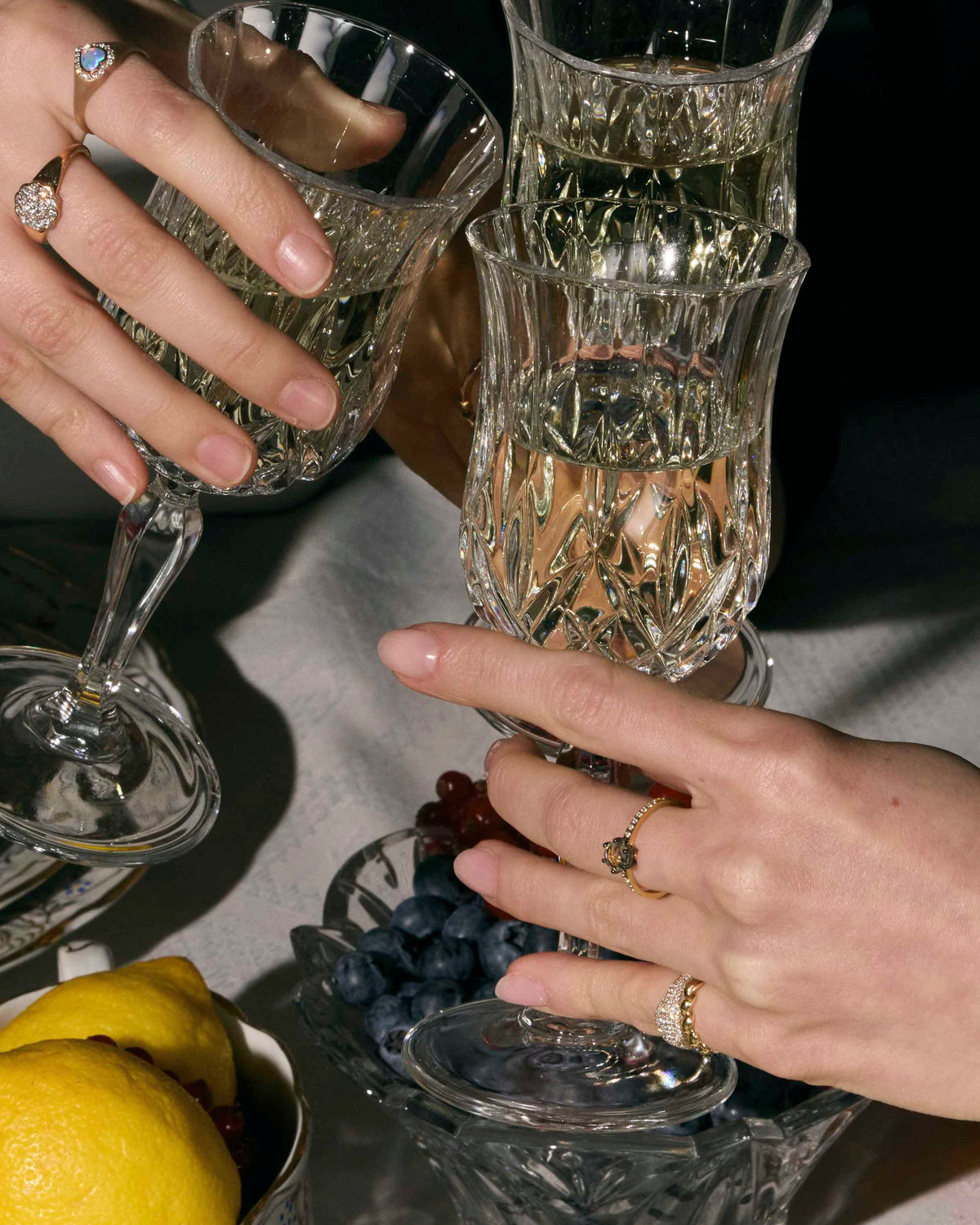 Two hands with gold rings with precious stones holding crystal glasses on a table with fruit and plates.