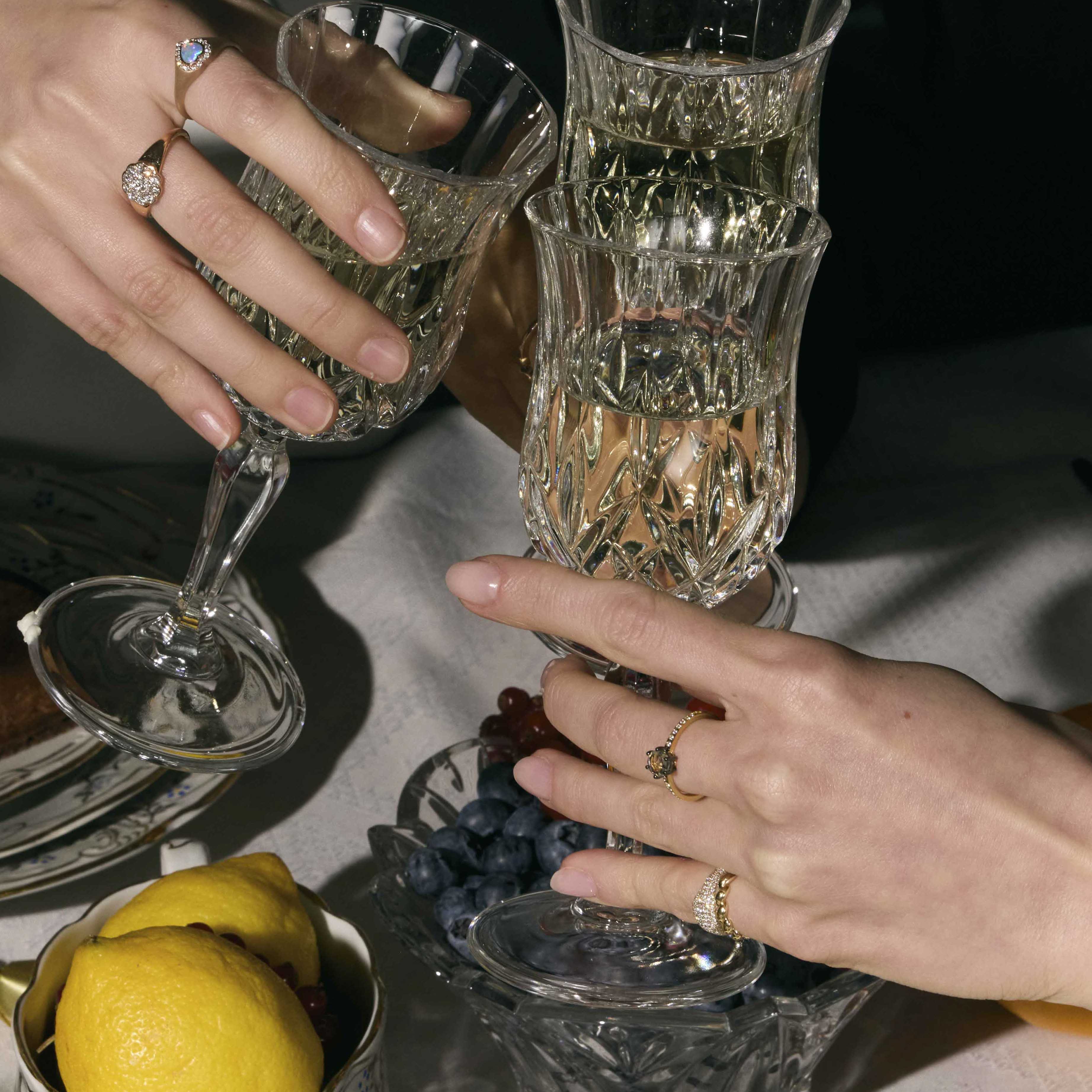 Two hands with gold rings with precious stones holding crystal glasses on a table with fruit and plates.