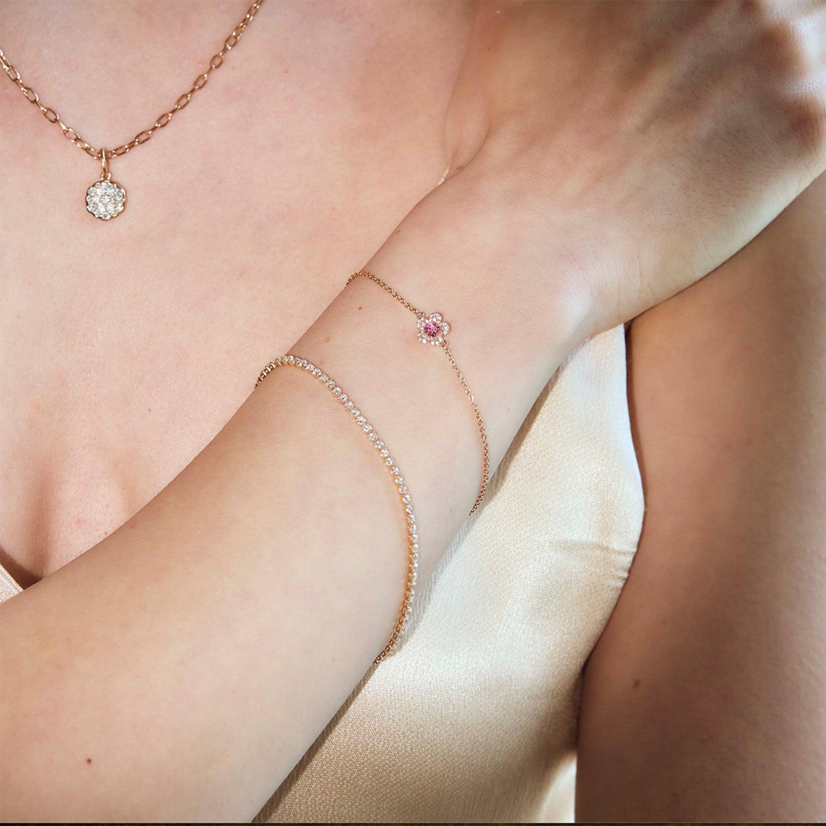 Close-up of a person wearing a delicate tennis gold bracelet with diamonds, a cluster diamonds pendant, a cherry blossom flower bracelet, on a neutral background.