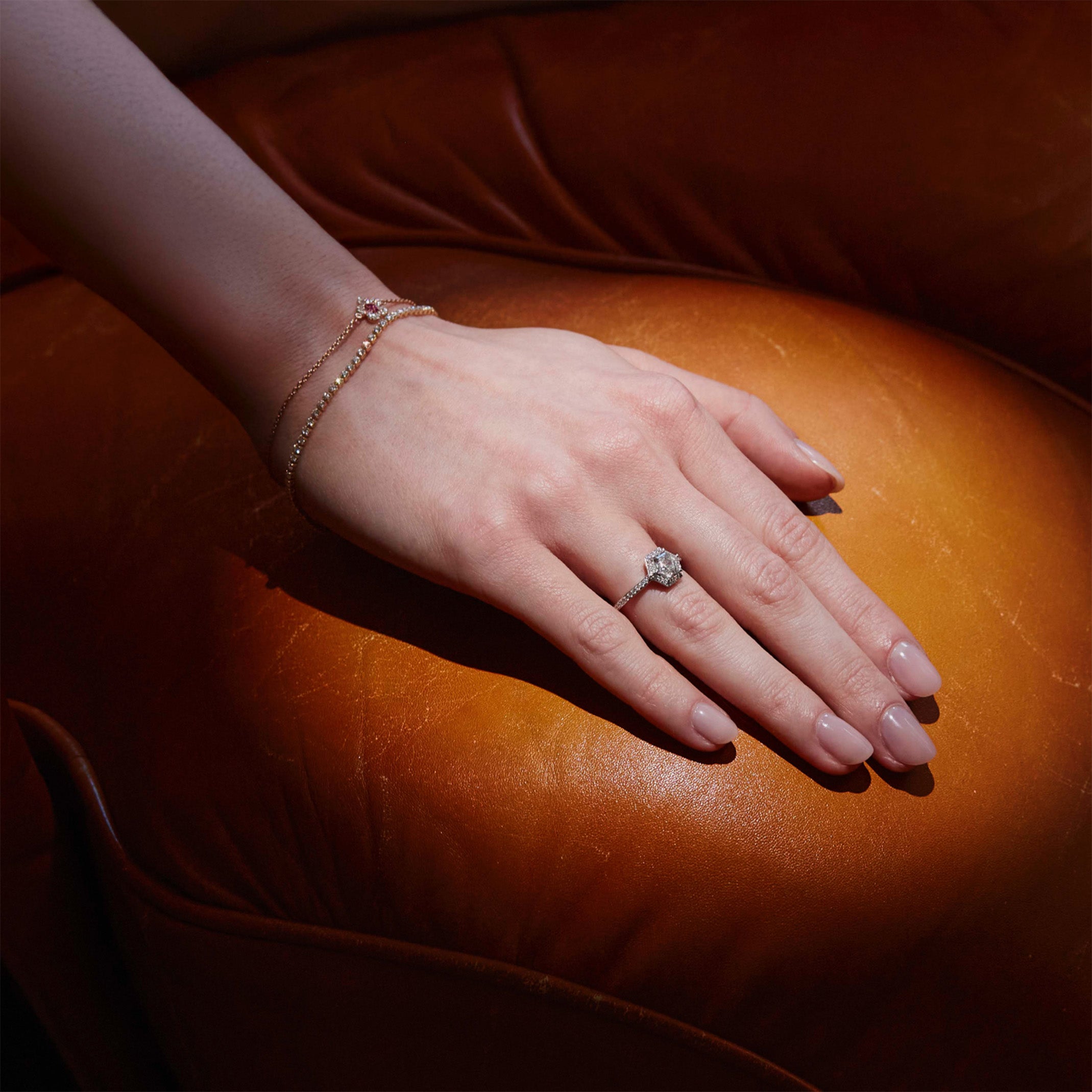 Hand wearing a hexagon diamond ring and two diamond bracelet on a brown leather couch