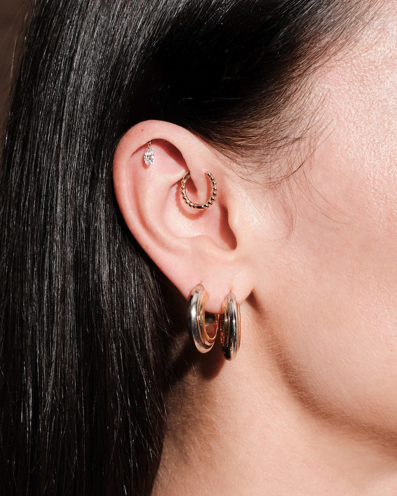 Close-up of an ear wearing multiple hoop earrings, thick gold hoops, floating marquise diamond stud, simple beaded hoop, with a blurred background