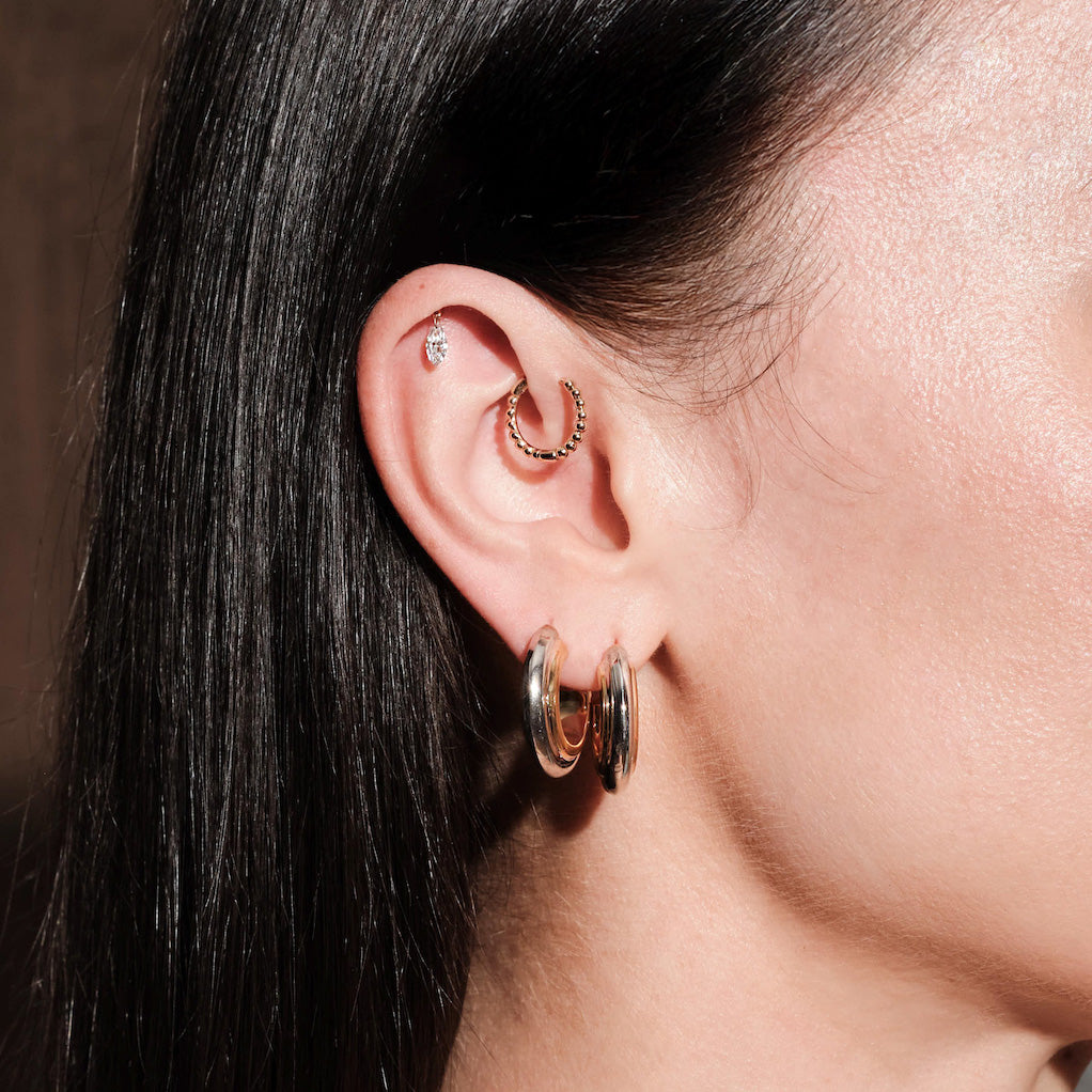 Close-up of an ear wearing multiple hoop earrings, thick gold hoops, floating marquise diamond stud, simple beaded hoop, with a blurred background