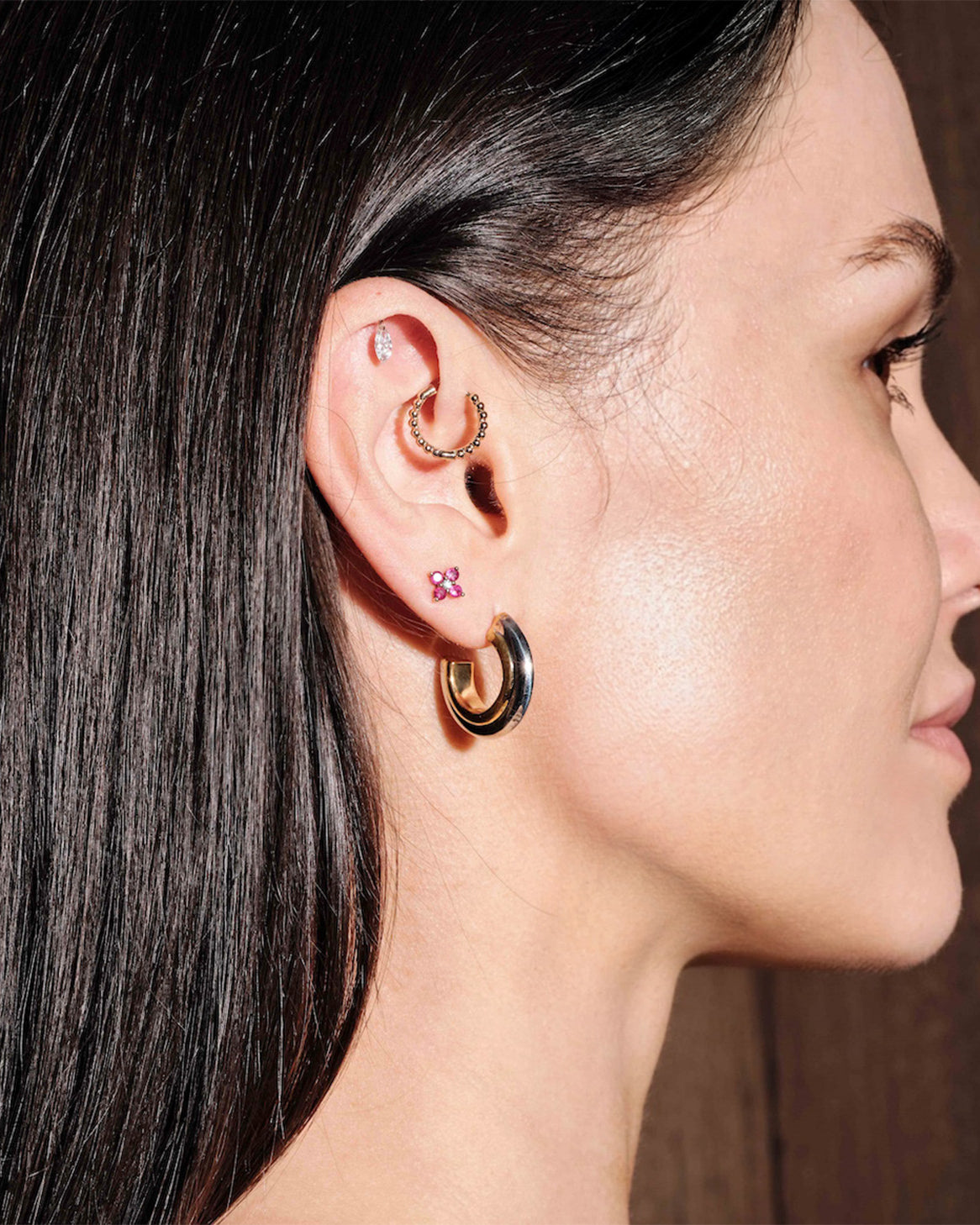 Woman wearing multiple hoop earrings, thick gold hoops, floating marquise diamond stud, simple beaded hoop, ruby gold studs, with a dark background