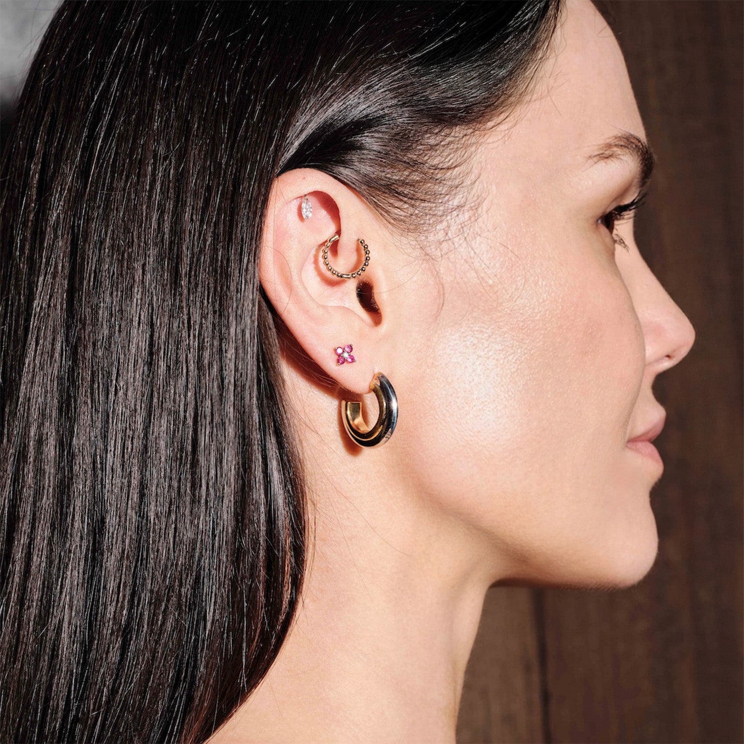 Woman wearing multiple hoop earrings, thick gold hoops, floating marquise diamond stud, simple beaded hoop, ruby gold studs, with a dark background