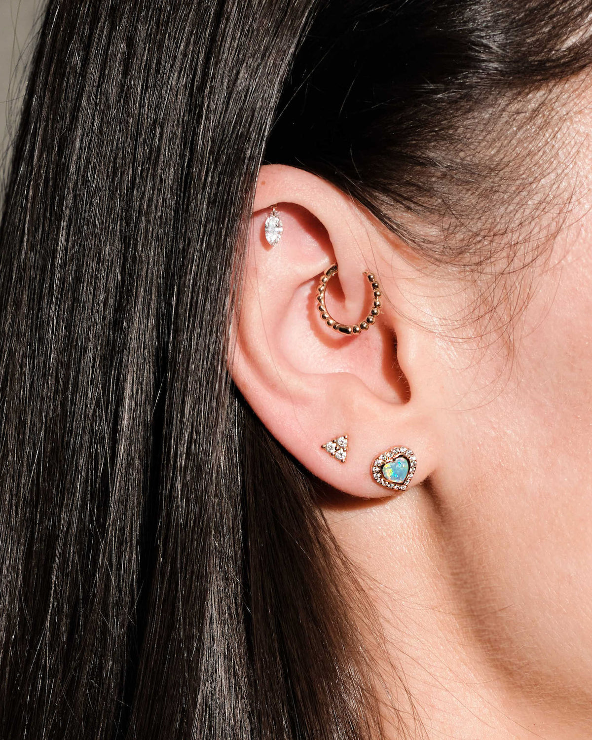 Close-up of an ear with multiple earrings, floating marquise diamond, beaded hoop, diamonds triangle stud, australian opal and diamonds heart studs, on a dark background