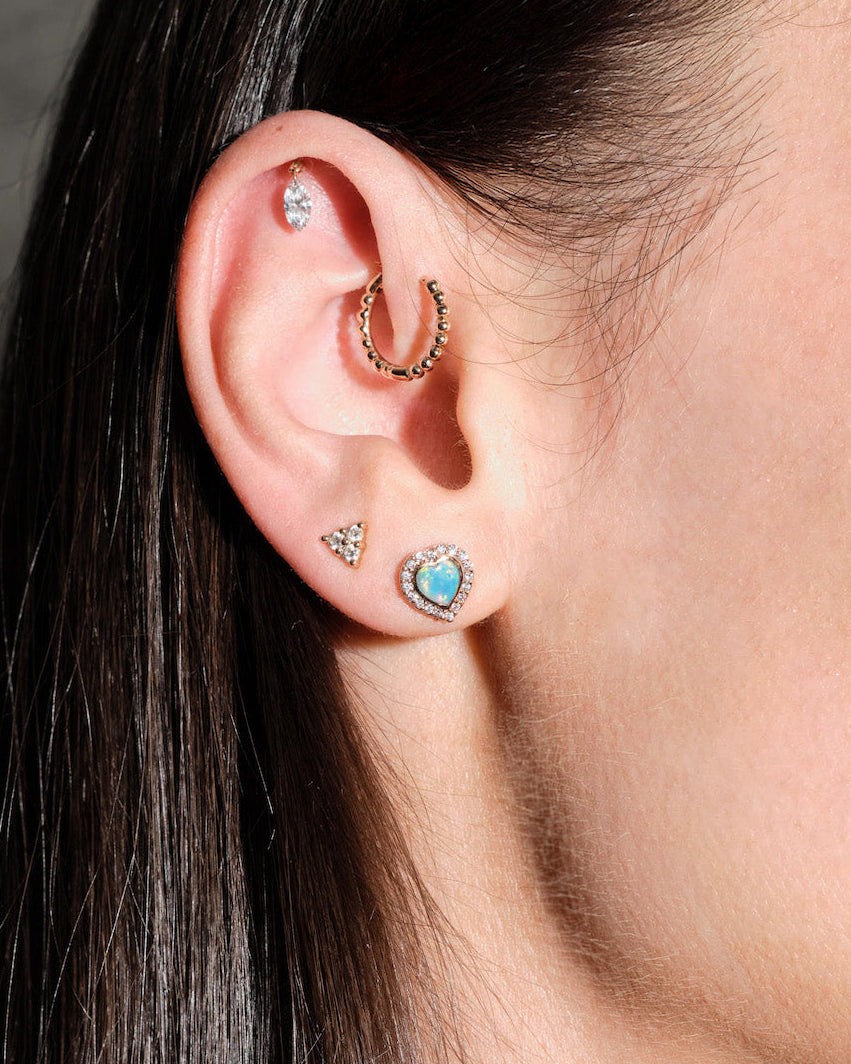 Close-up of an ear with multiple earrings, floating marquise diamond, beaded hoop, diamonds triangle stud, australian opal and diamonds heart studs, on a dark background