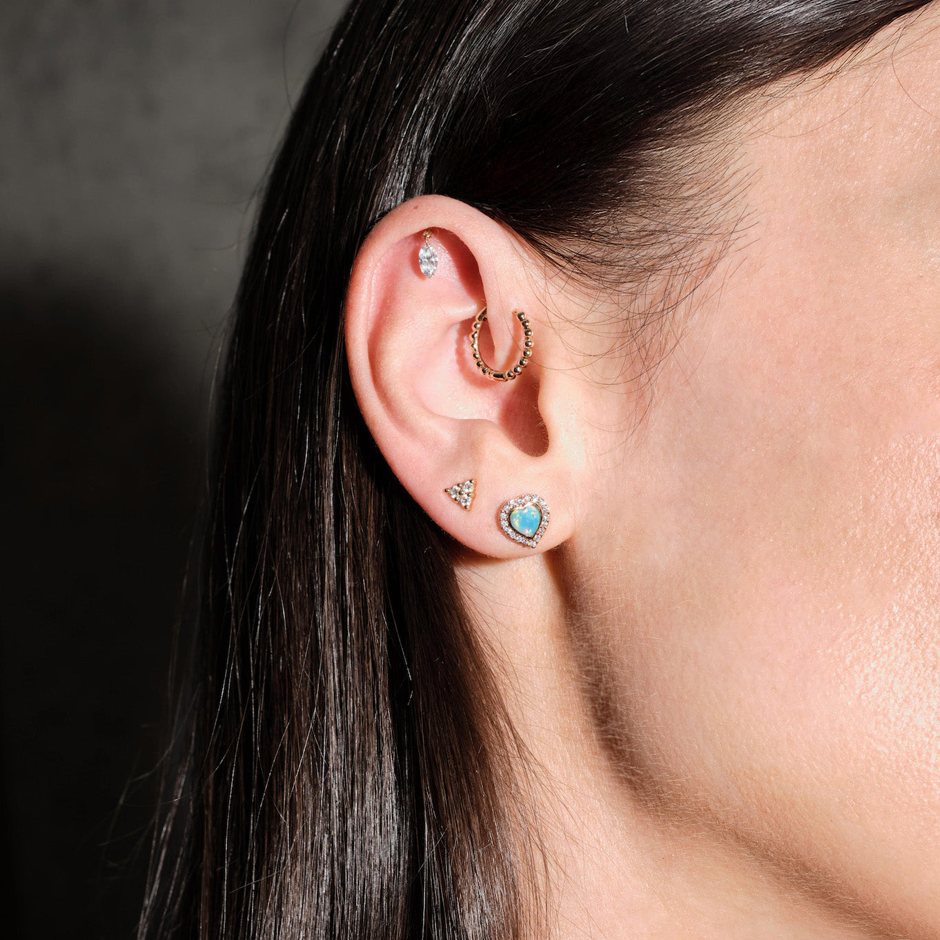 Close-up of an ear with multiple earrings, floating marquise diamond, beaded hoop, diamonds triangle stud, australian opal and diamonds heart studs, on a dark background