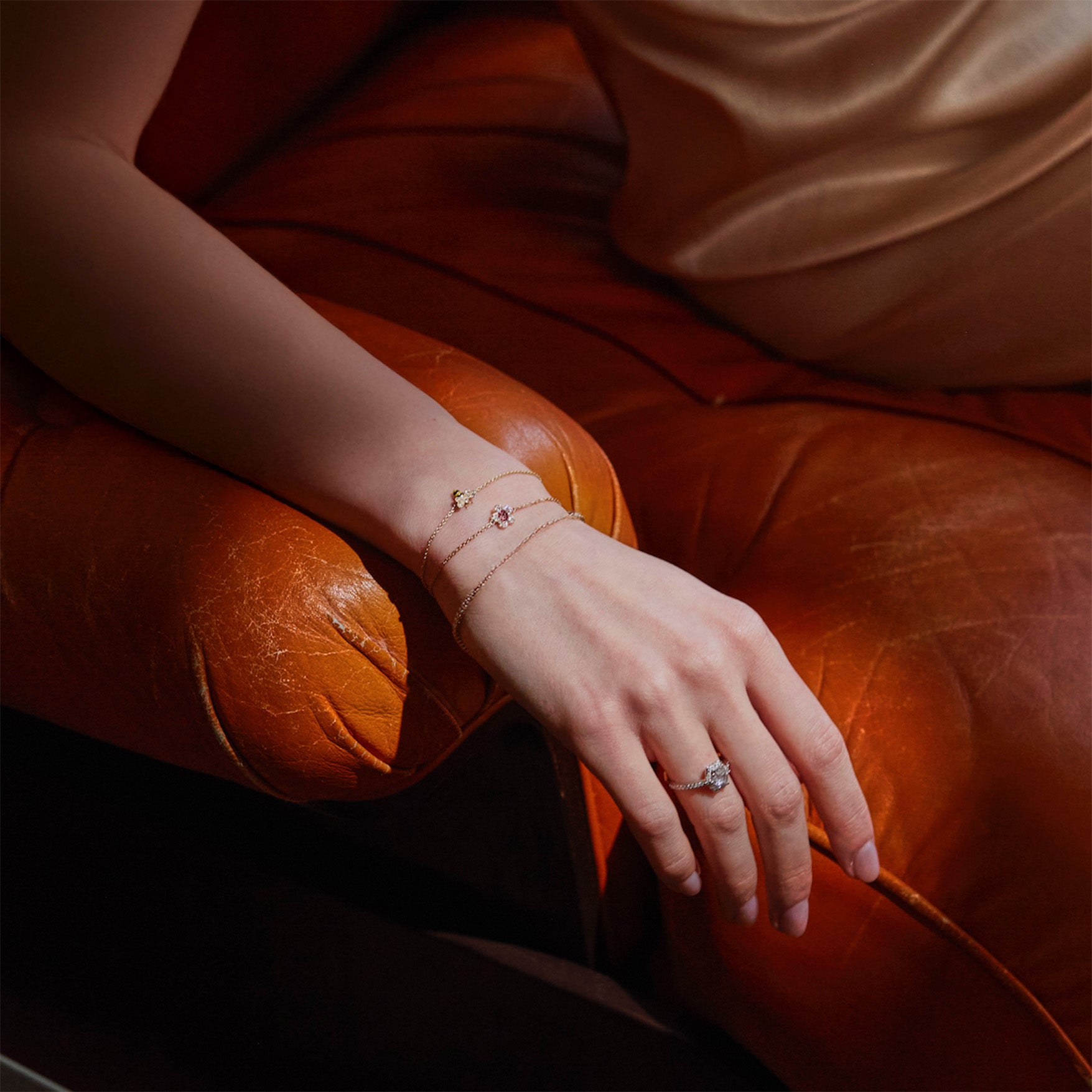 Close-up of a hand wearing gold bracelets, a honey bee bracelet, a cherry blossom flower bracelet and a tennis diamonds bracelet, on a brown leather couch