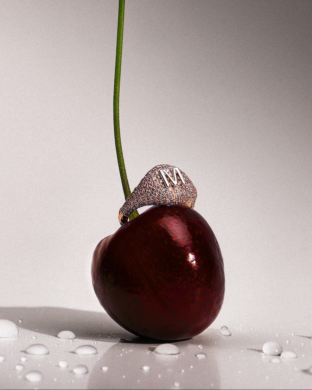 A dark red cherry with a long green stem stands upright on a glossy white surface with water droplets. A pave diamonds signet 14k gold ring with a large white gold letter 'M' sits on the cherry's stem.