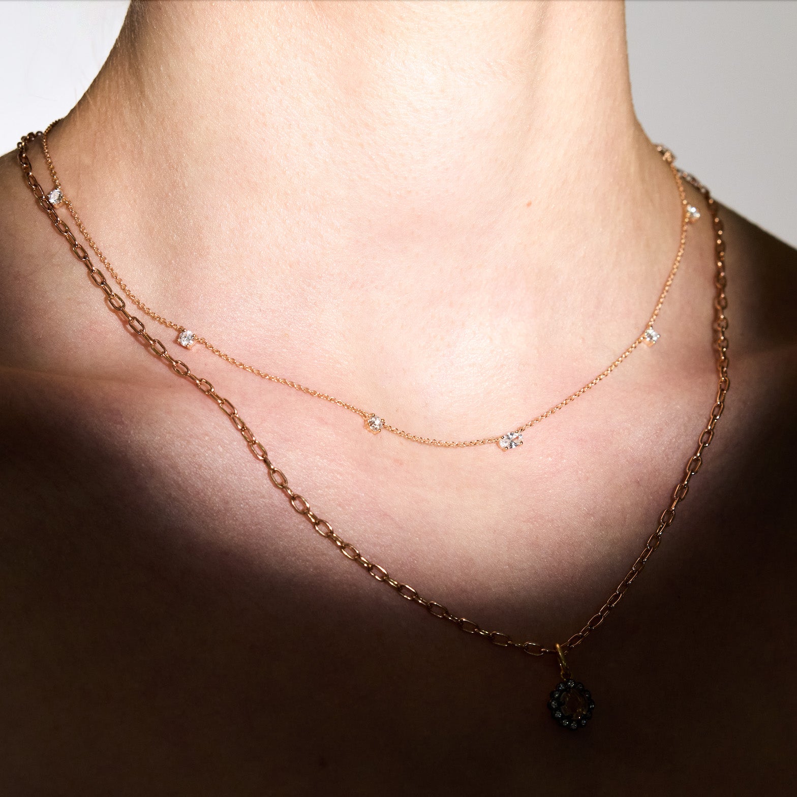 Two gold necklaces with, one with small diamonds scattered on a chain, on a person's neck against a neutral background