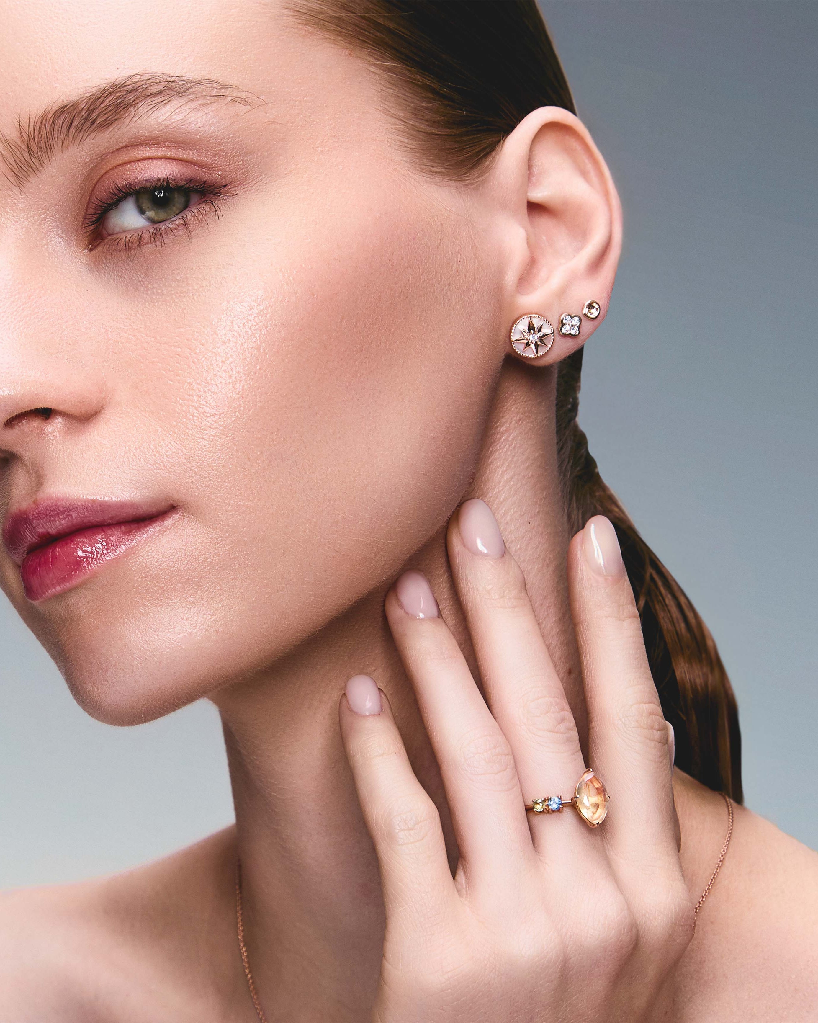 A pair of gold studs earrings with a simple design, resembling a prayer bowl or birds' nest, a clover diamonds earring, a mother of pearl lodestar earring, on an ear.