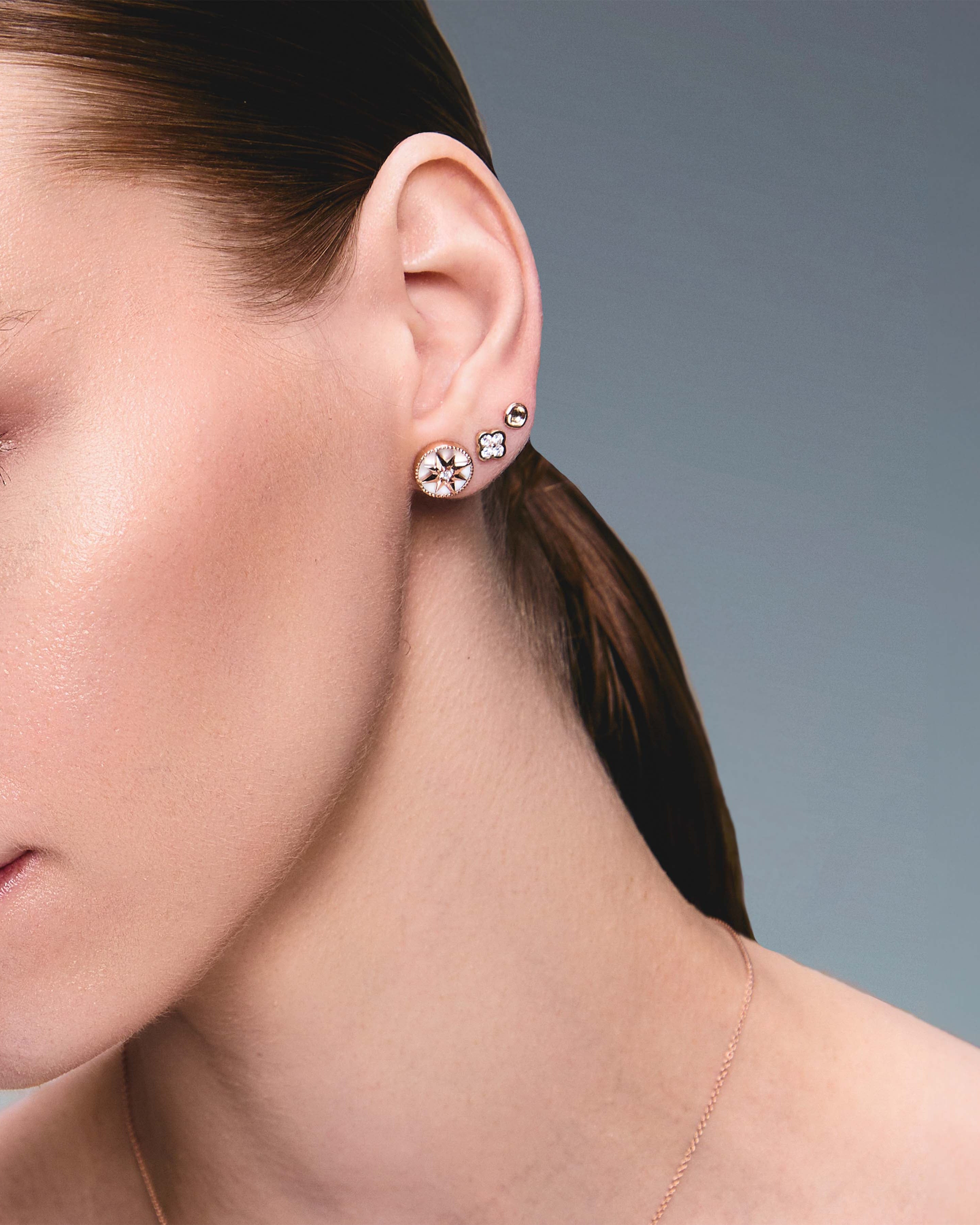 A pair of gold studs earrings with a simple design, resembling a prayer bowl or birds' nest, a clover diamonds earring, a mother of pearl lodestar earring, on an ear.