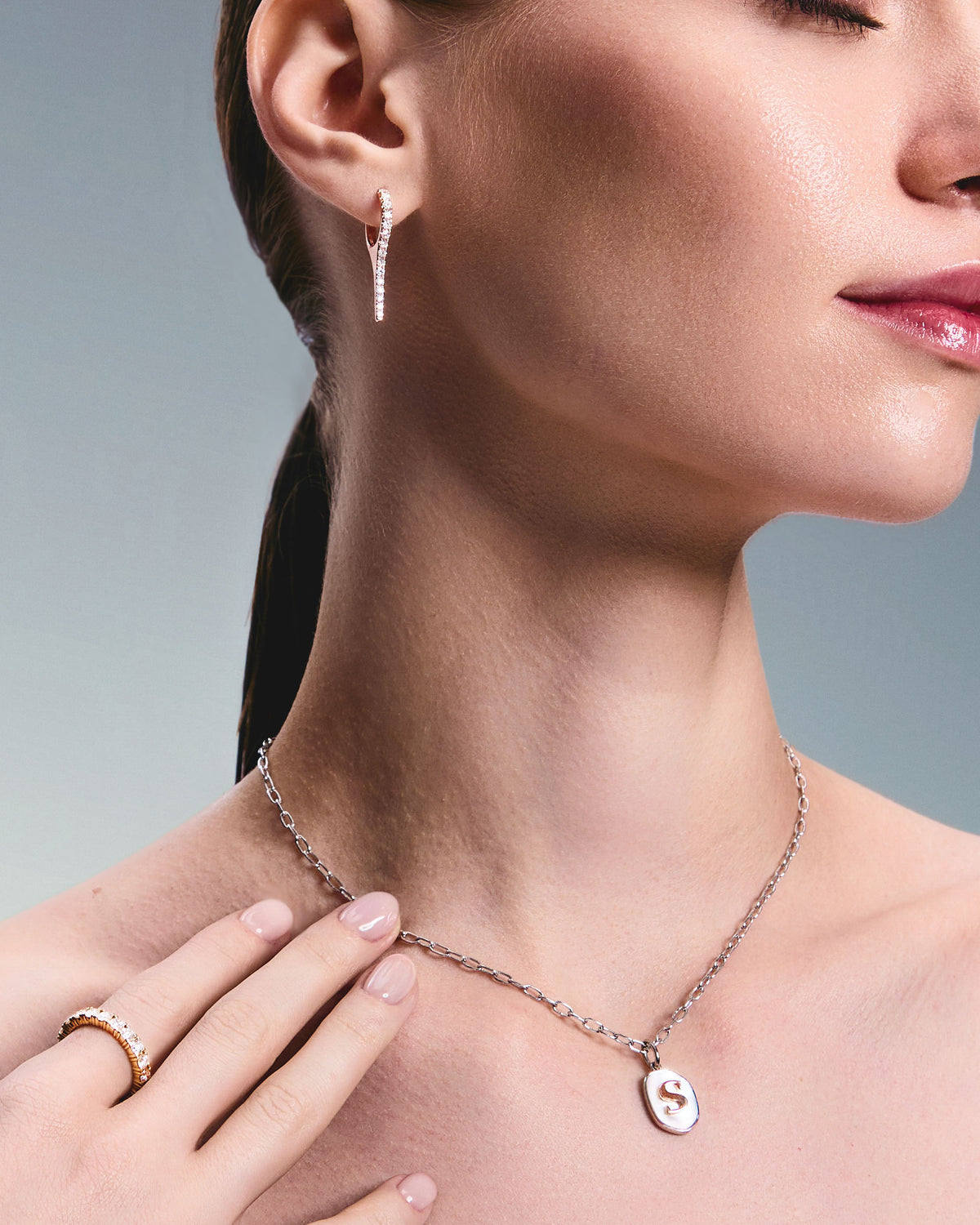 A person wearing a necklace with a gold initial pendant made of Mother of Pearl and a pair of rose gold earrings with natural diamond drops, featuring a minimalist design.