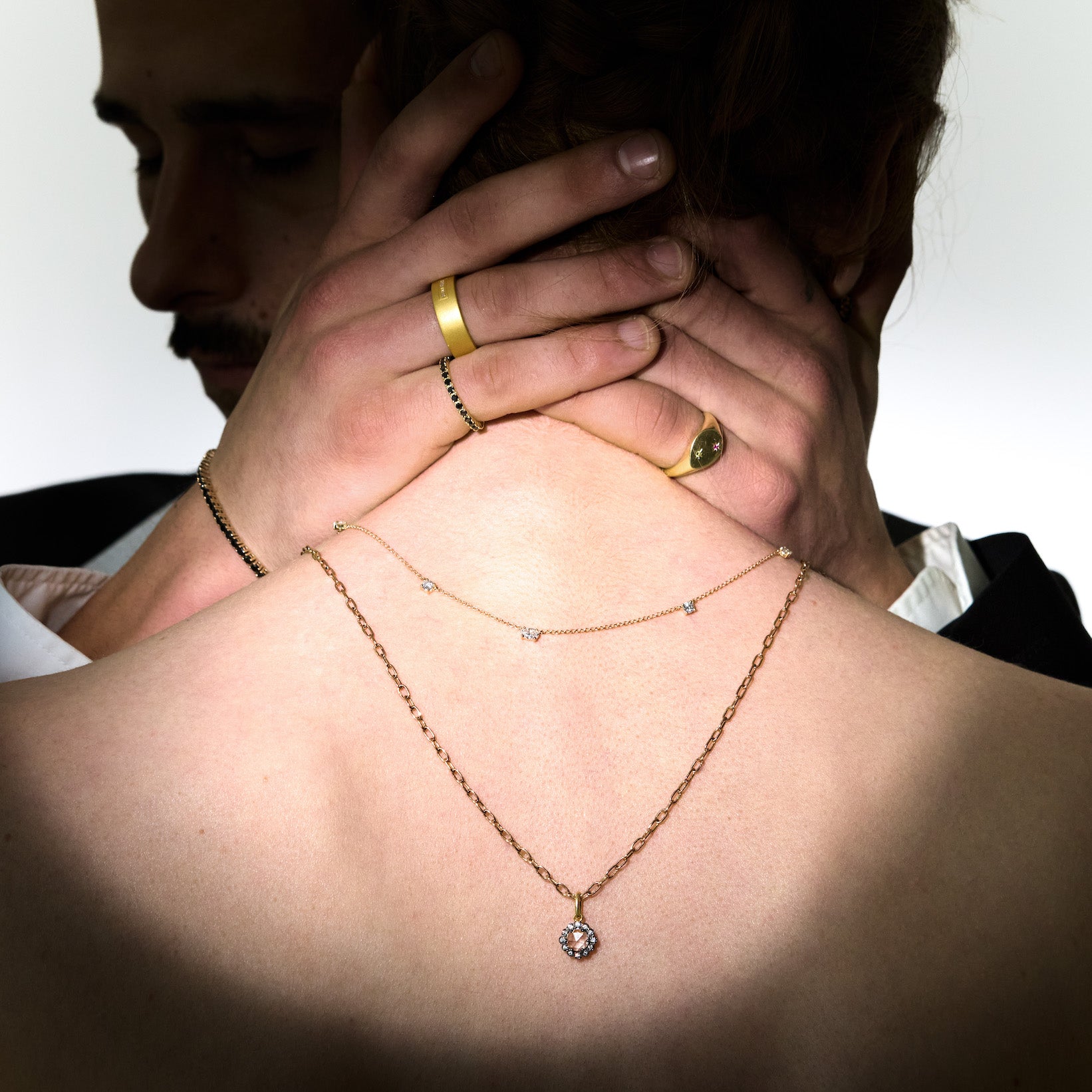 a couple hugging, she is wearing two gold necklaces and he is fashioning multiple rings on a plain background