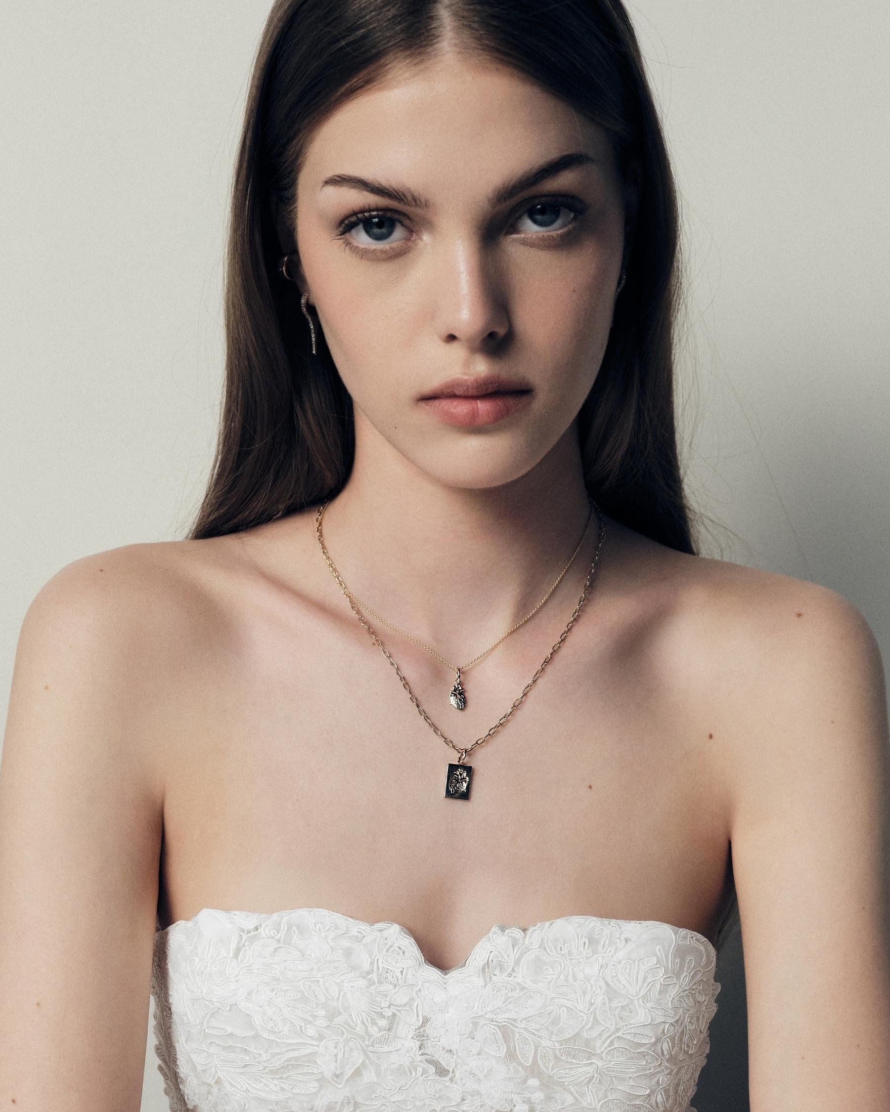 Woman wearing a strapless white lace dress with jewelry, a necklace with 14k gold heart plates on a neutral background