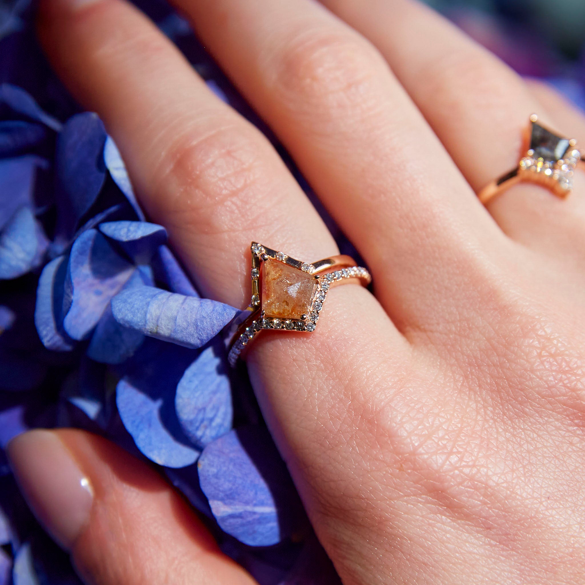 A hand wearing a gold band ring with a large, square-cut orange diamond surrounded by smaller white diamonds on the band, with a background of blue flowers.