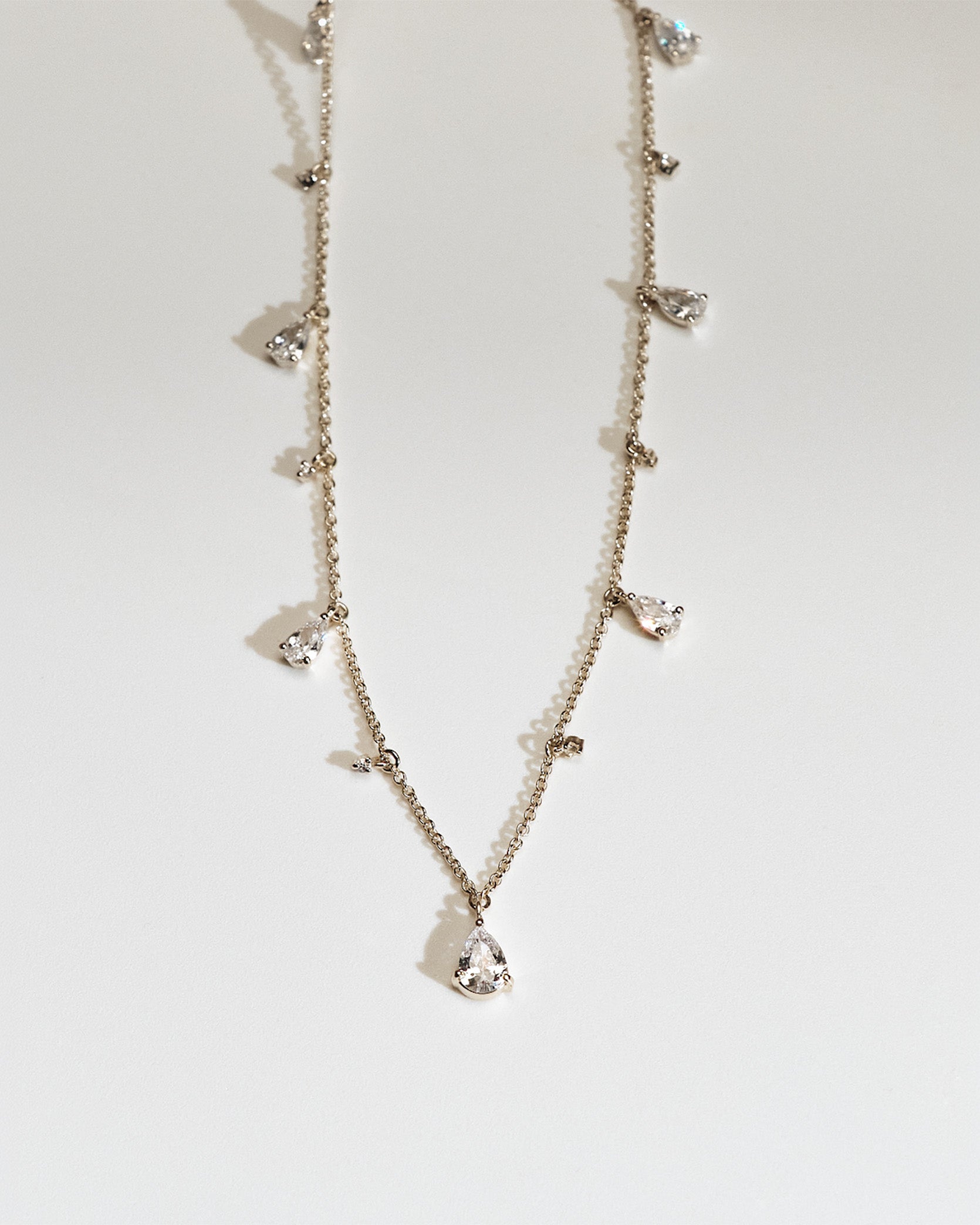 Delicate gold necklace with small teardrop and round shaped diamond pendants on a light gray background