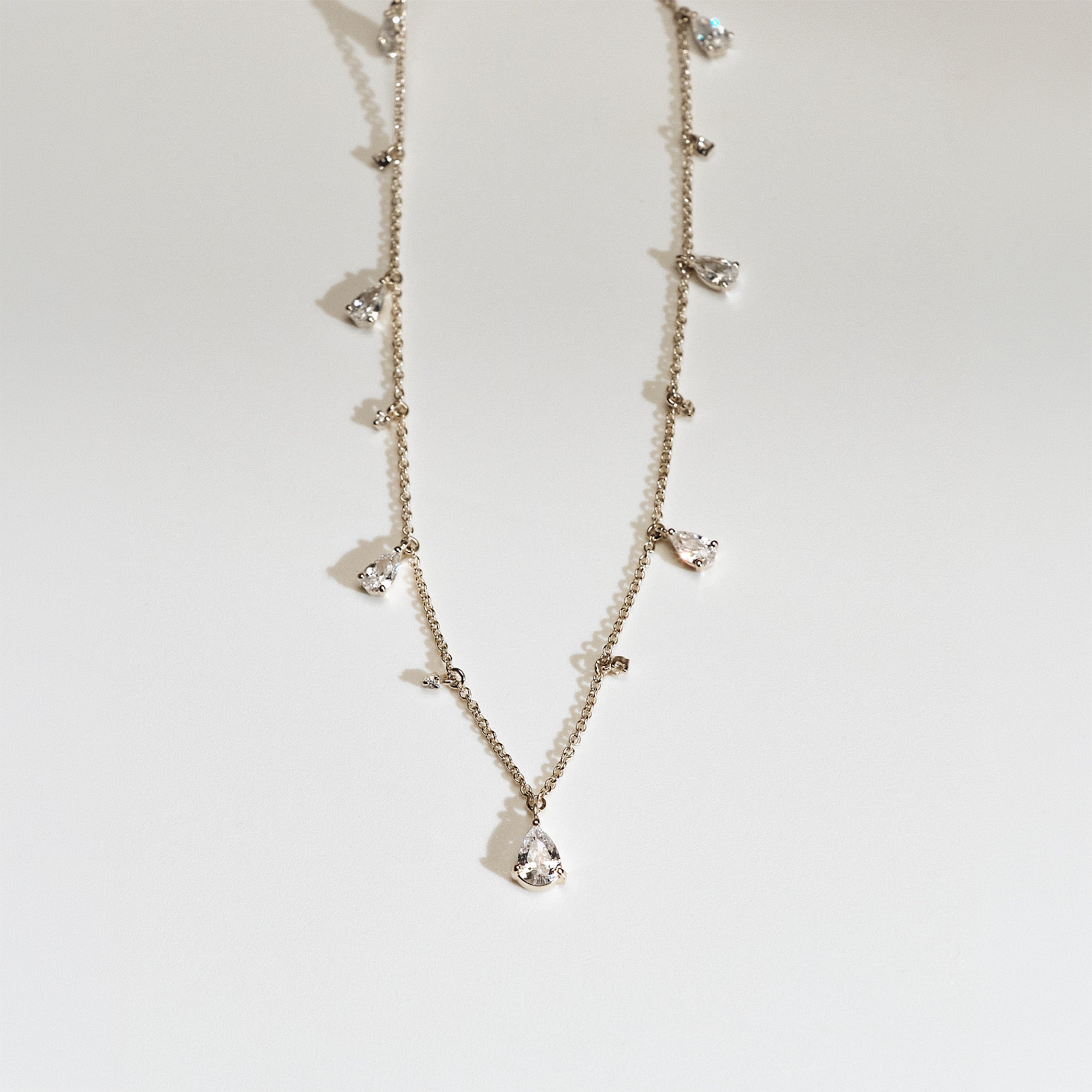 Delicate gold necklace with small teardrop and round shaped diamond pendants on a light gray background