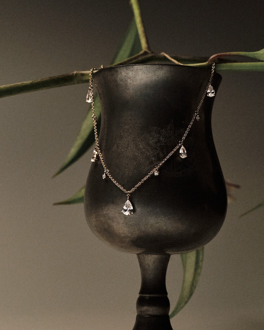 Gold Necklace with teardrop and round diamonds pendants on a black stand with green leaves in the background.