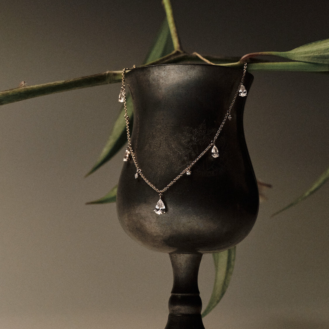 Gold Necklace with teardrop and round diamonds pendants on a black stand with green leaves in the background.