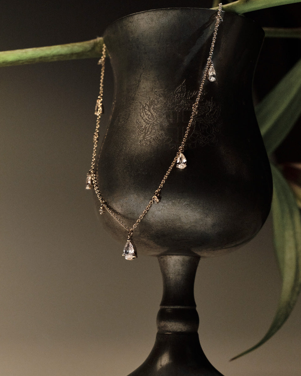 Gold Necklace with teardrop and round diamonds pendants on a black stand with green leaves in the background.