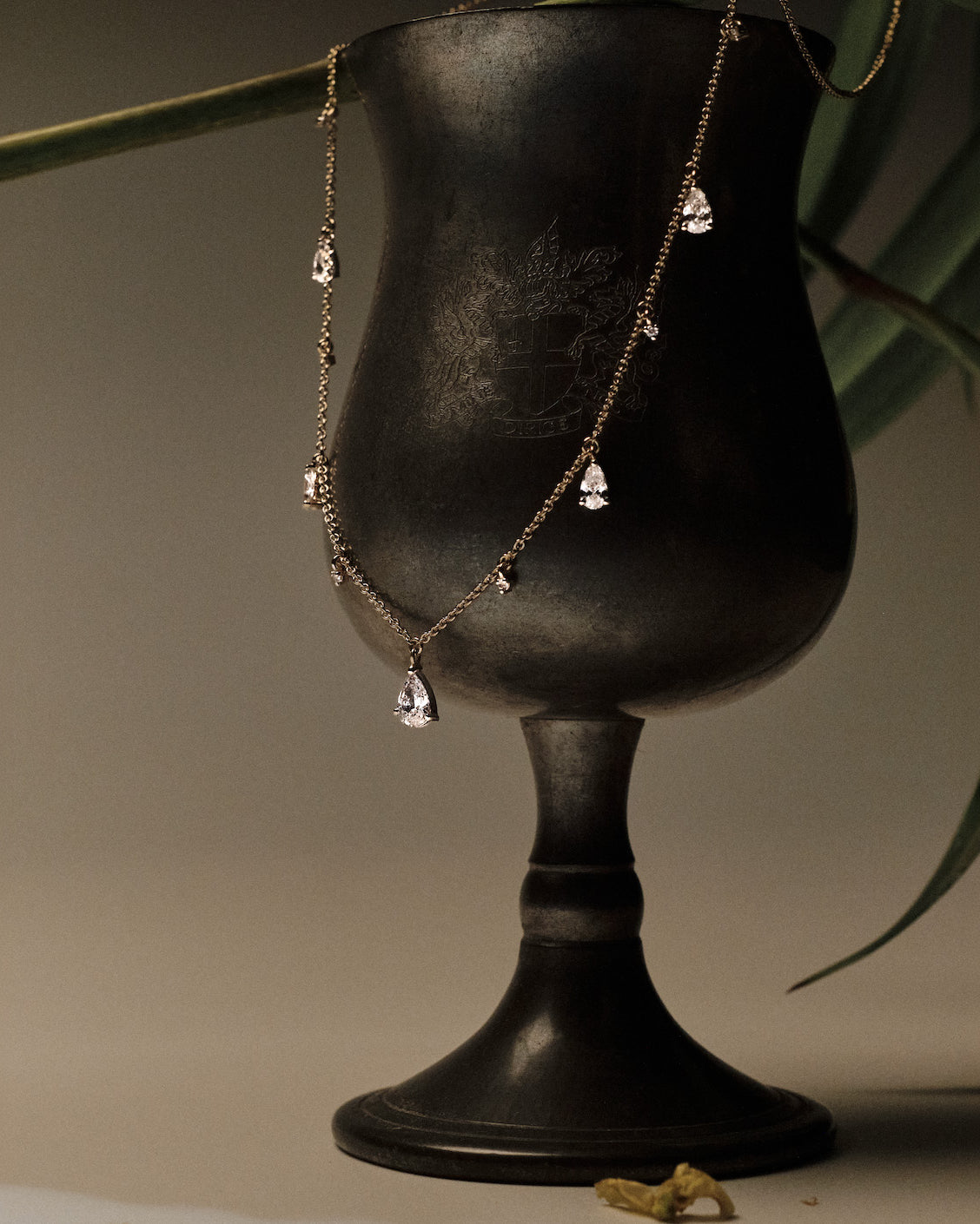 Gold Necklace with teardrop and round diamonds pendants on a black stand with green leaves in the background.