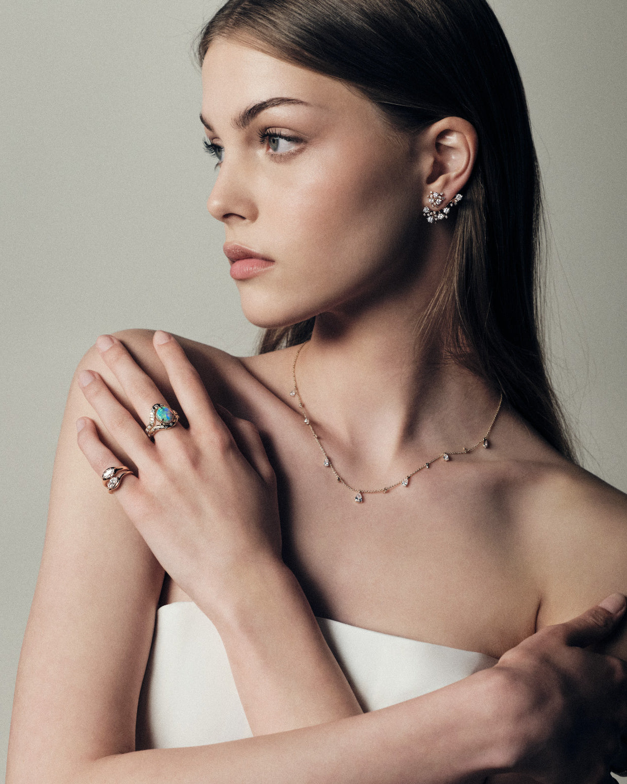 Woman wearing gold jewelry including bloom diamonds earrings, raindrops teardrops necklace, marquise diamonds signet pinky rings, and an australian opal and diamonds ring on a neutral background.