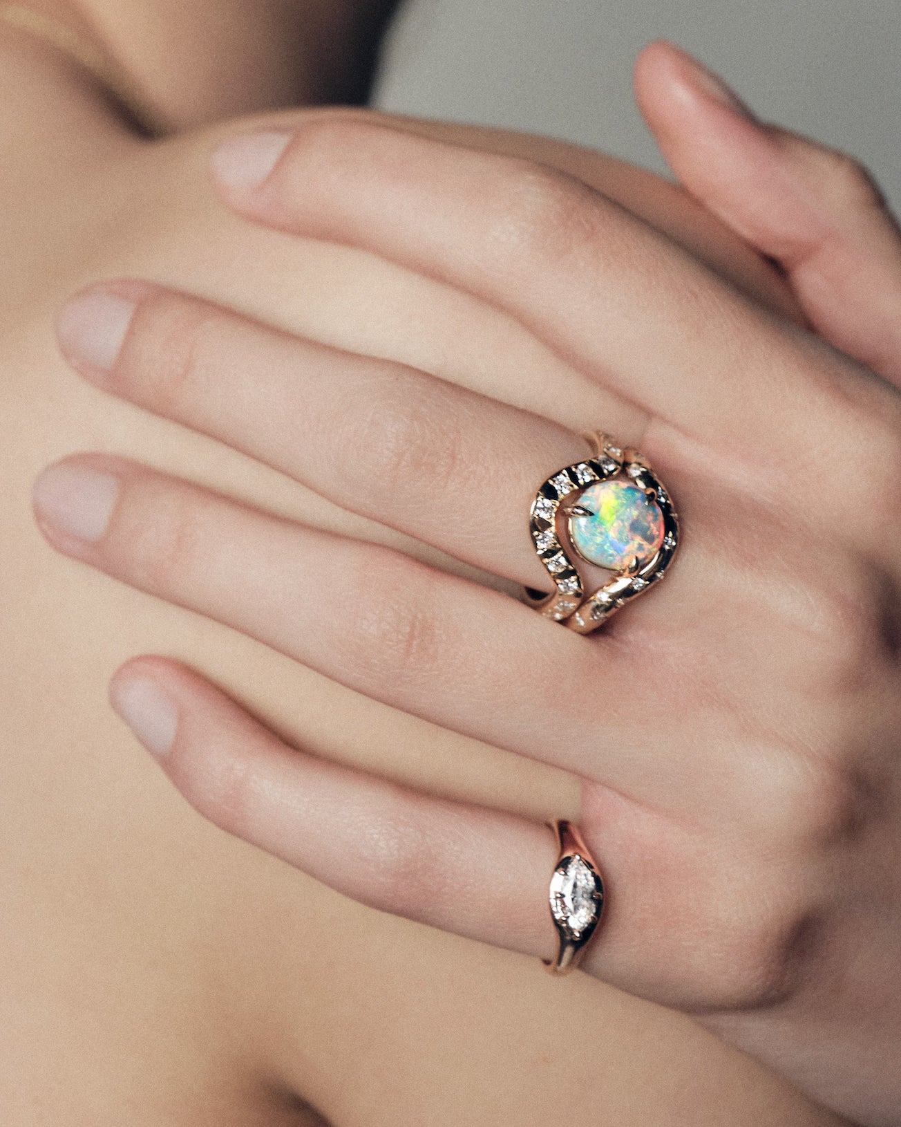 Close-up of a hand wearing a marquise diamond signet ring, and a band with an australian opal ring on a neutral background.