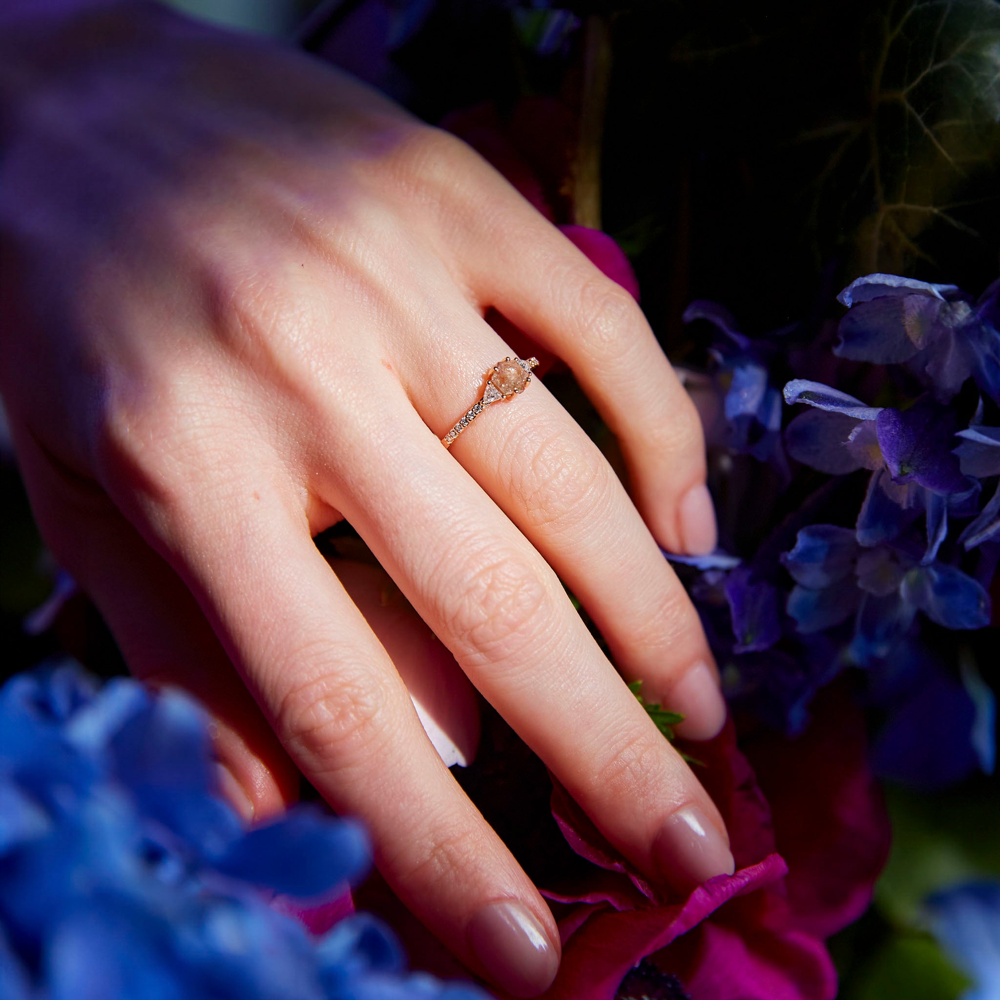 Hexagon Salt and Pepper diamond ring on hand surrounded by flowers. Inel cu diamant hexagon salt and pepper pe mână, încunjurată de flori.