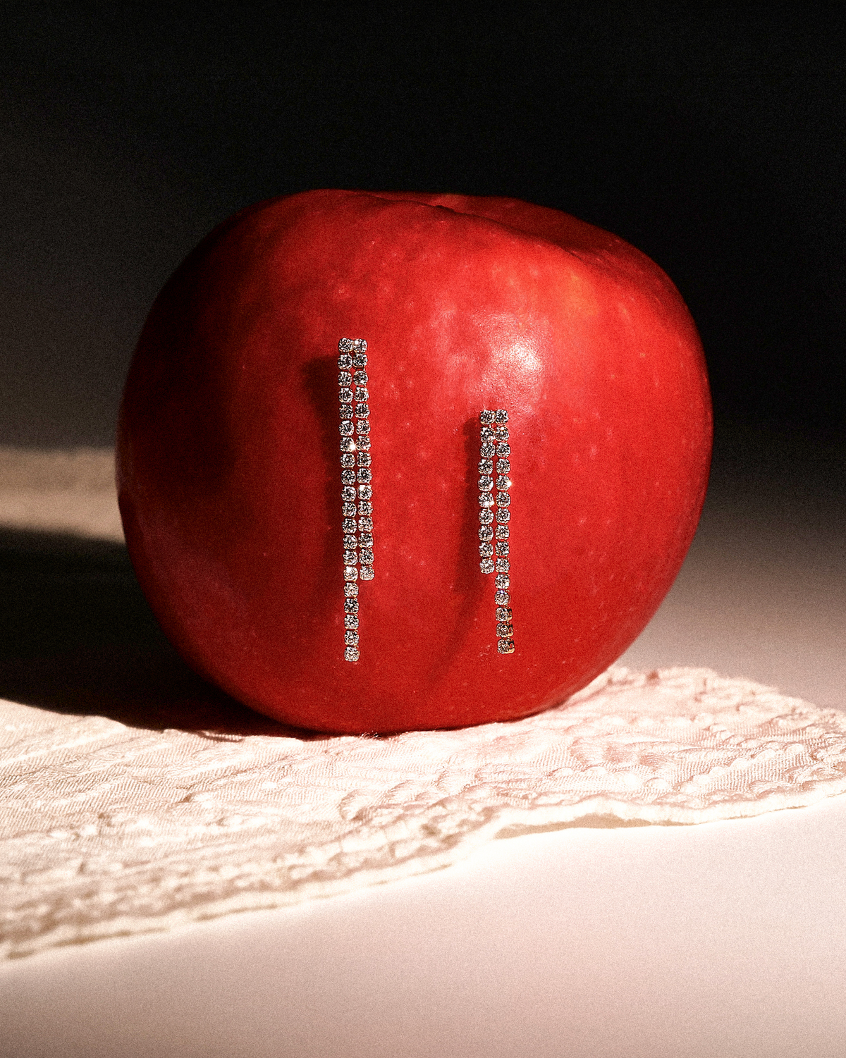A red apple with two vertical rows of sparkling white gold earrings attached to its side, photographed on a white surface with a lace cloth and a dark background.