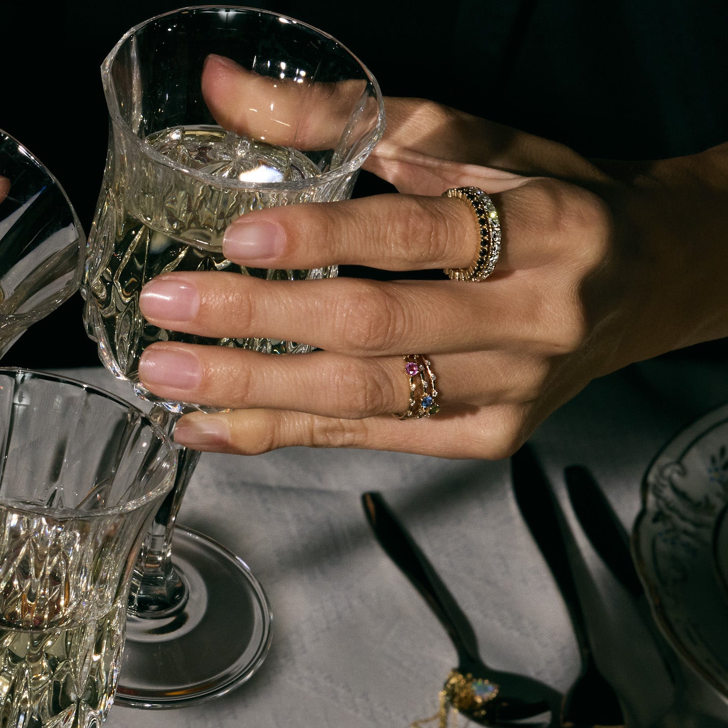 Hand full of rings, eternity black and white diamond rings, holding a crystal glass with another glass in the foreground, on a dark background