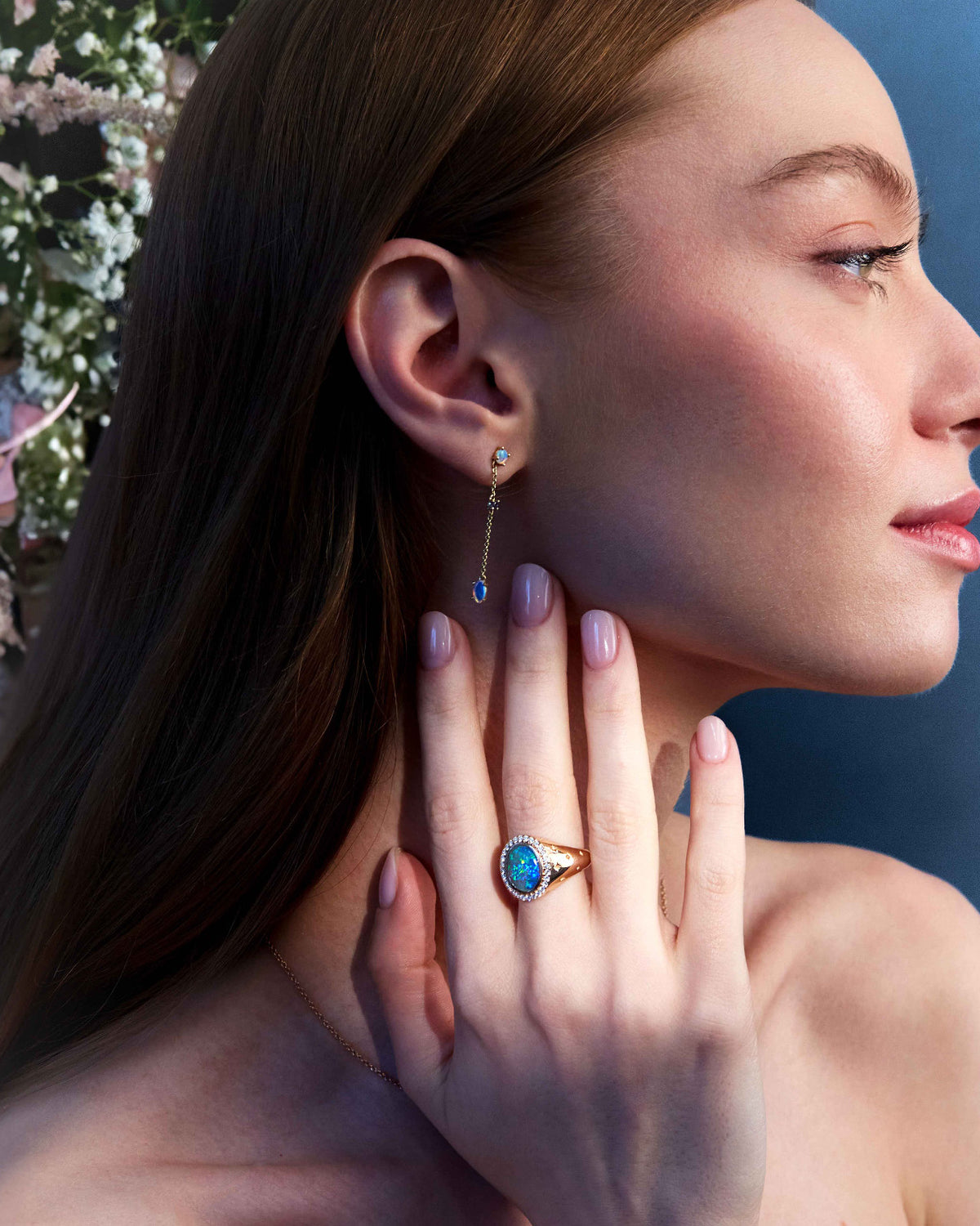 A person wearing a pair of gold earrings with round blue or green sapphires and Australian Opals, and an Australian Oval Opal Signet ring, standing near a floral backdrop.
