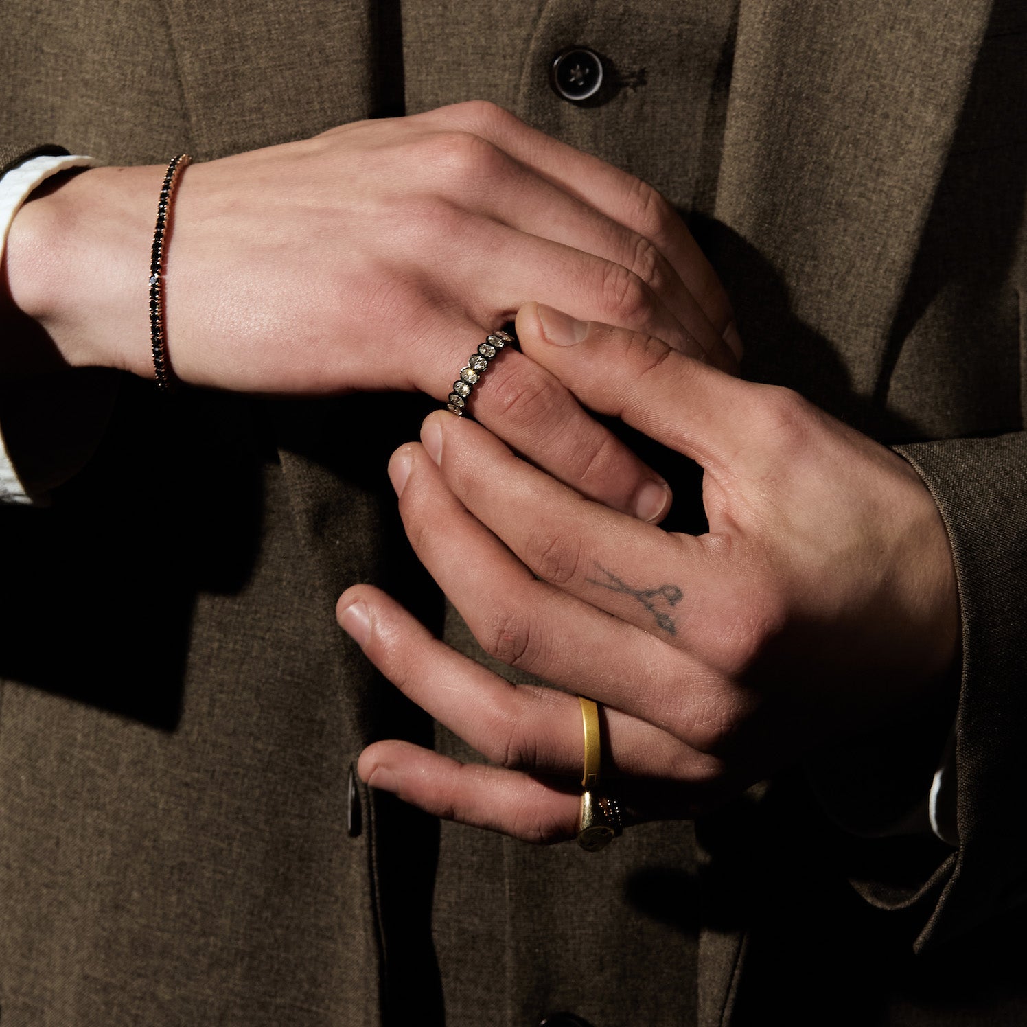 Close-up of two hands with rings - eternity diamonds and black gold ring, and wearing a black diamonds tennis bracelet, against a brown background.