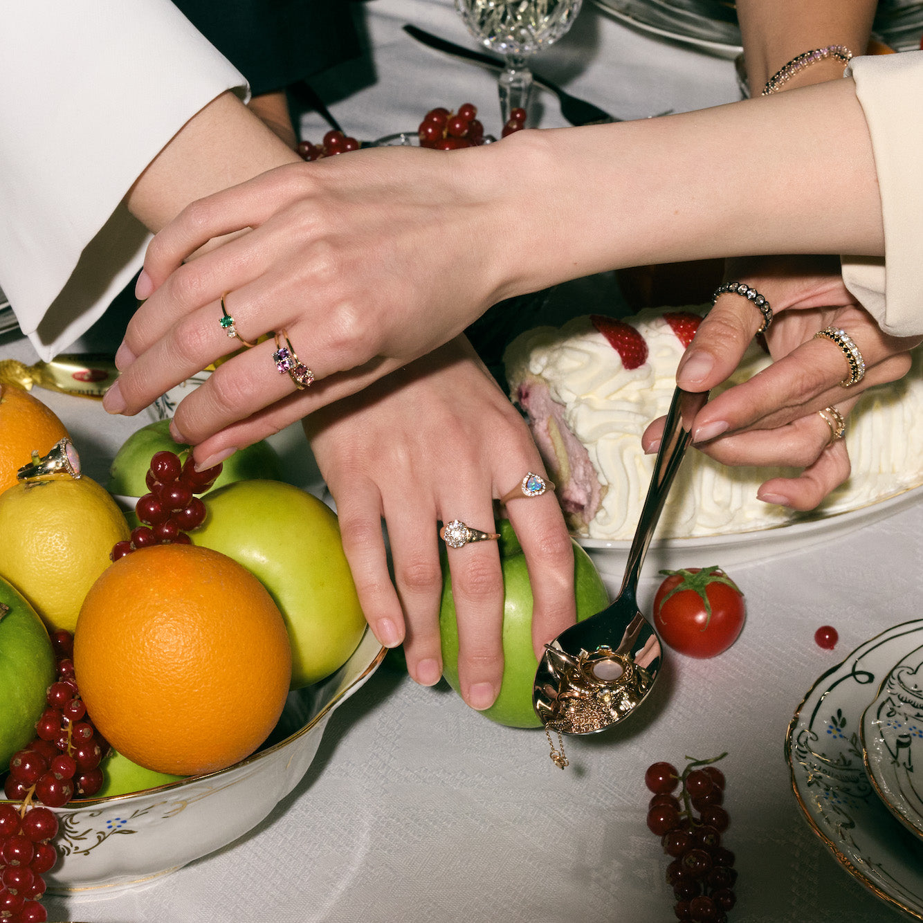 Close-up of hands with gold rings around a table with fruit and cake.