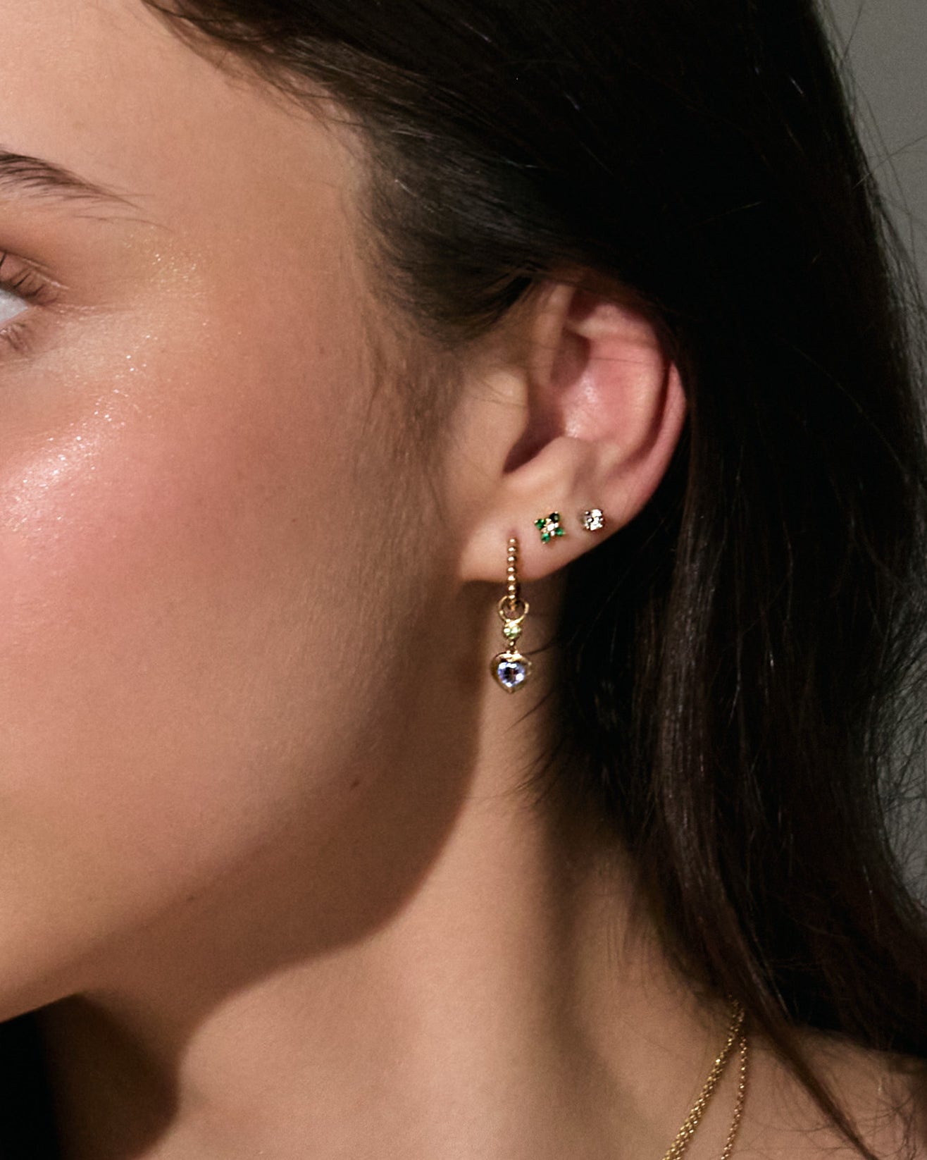 Close-up of a woman wearing stud 0,20cts diamond earrings, emerald earrings and hoop earrings with a neutral background
